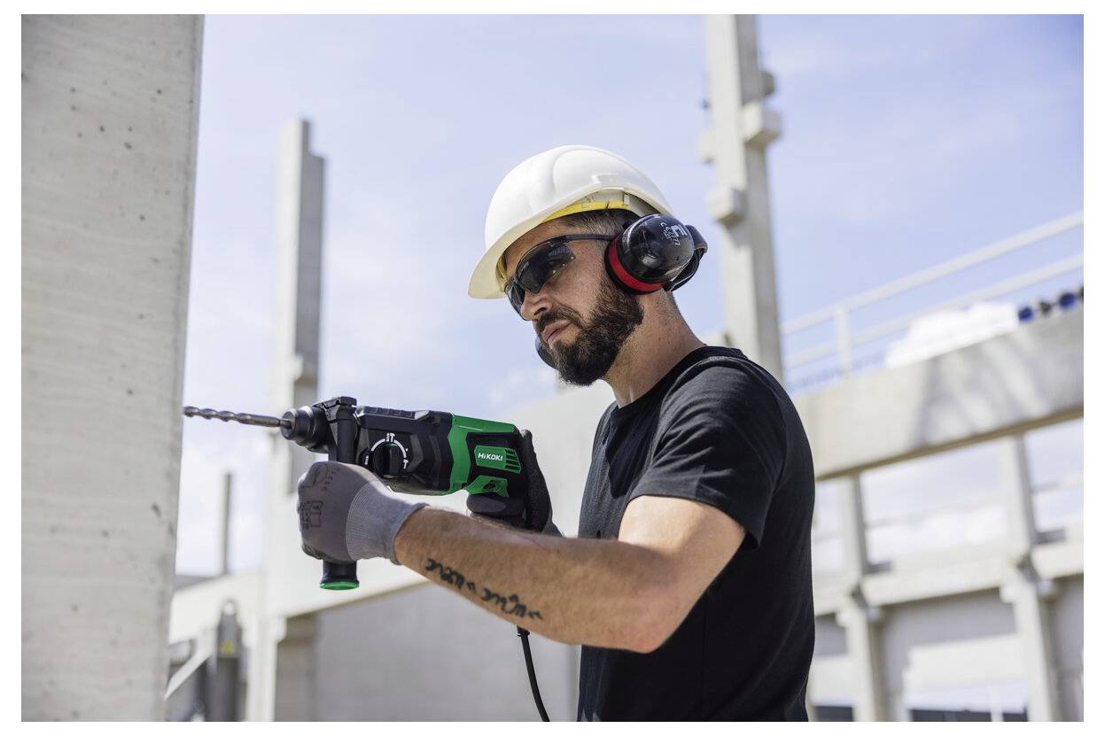 Bauarbeiter mit Schutzhelm und Gehörschutz bohrt mit einem Bohrer in eine Betonwand auf einer Baustelle.