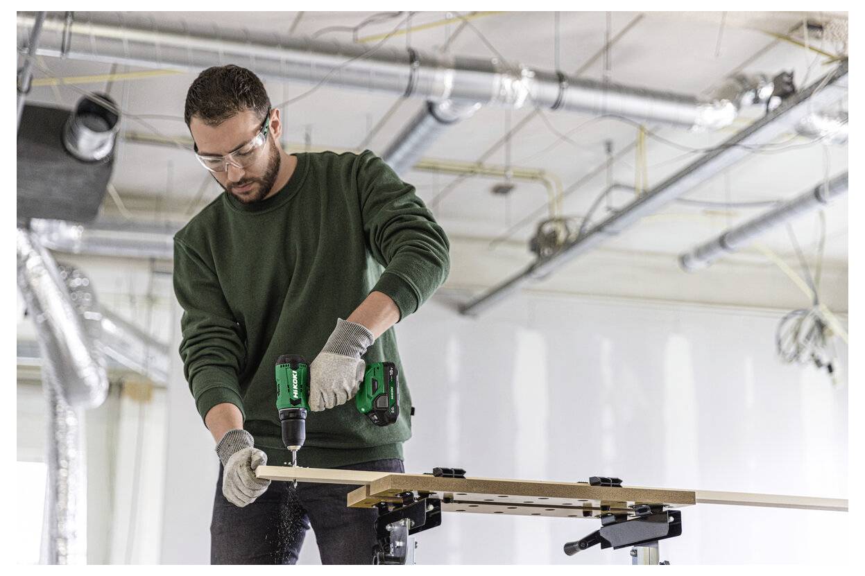 Ein Mann trägt eine Schutzbrille und Handschuhe und verwendet einen Akku-Bohrer, um ein langes Holzbrett in einer Baustelle unter Konstruktion zu befestigen.