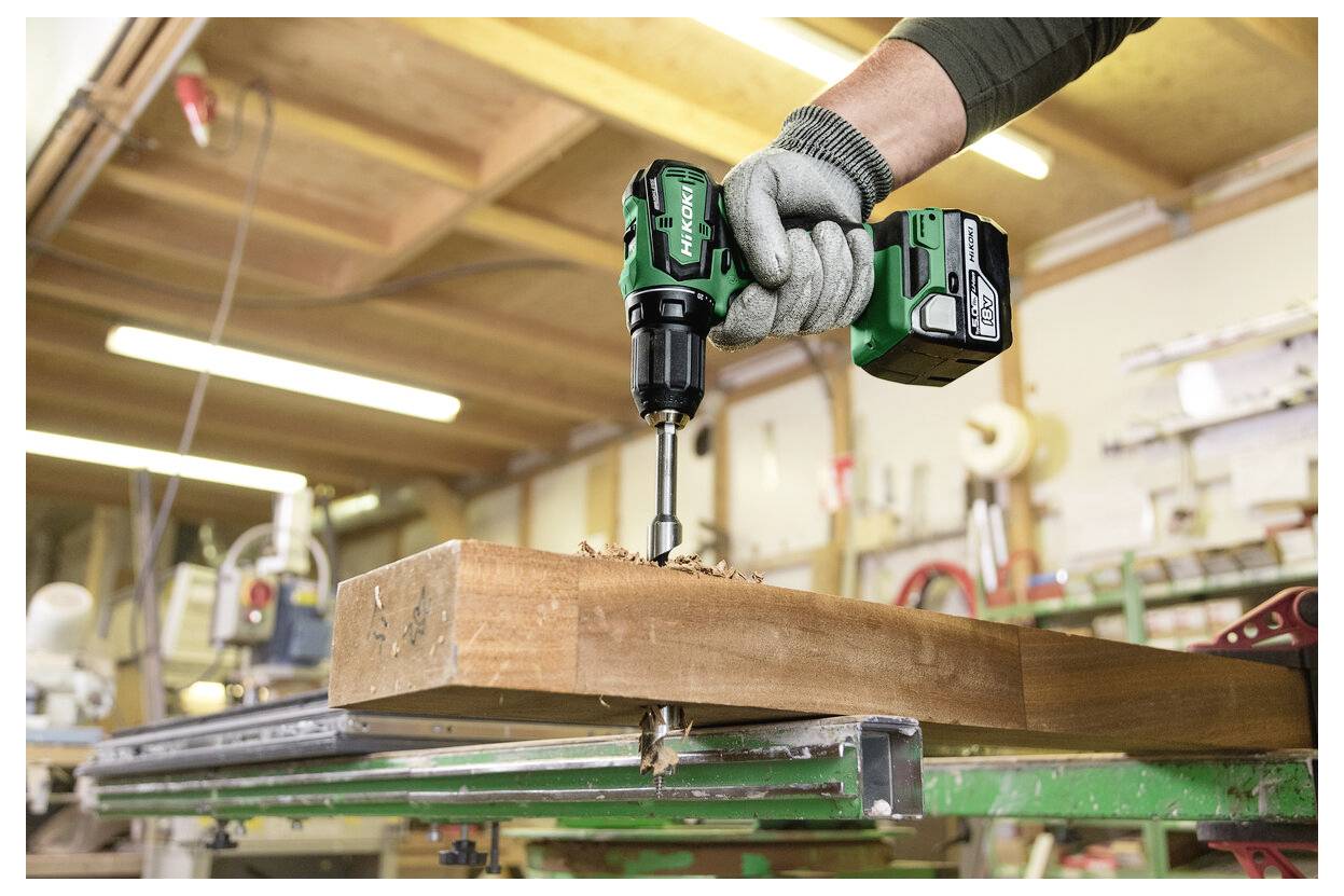 Ein Arbeiter mit Handschuhen nutzt einen kabellosen Bohrer, um ein Loch in einen Holzbalken in einer Werkstatt zu bohren.