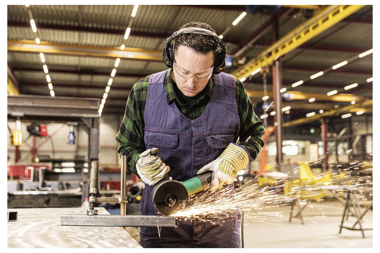 Ein Arbeiter in einem Lagerhaus schneidet Metall mit einem Winkelschleifer und sendet dabei Funken über die Werkstatt aus, während er eine Schutzbrille, Gehörschutz und Handschuhe trägt.