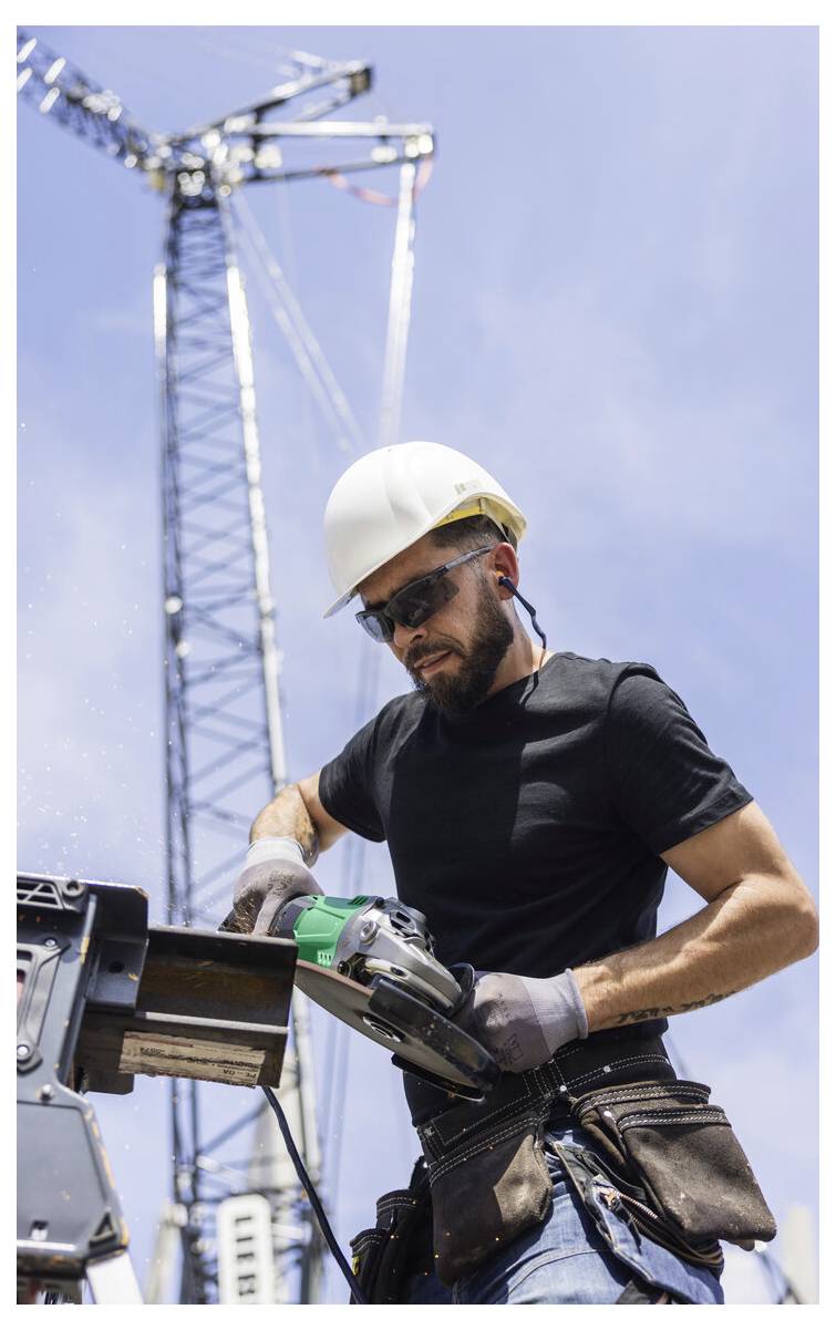 Bauarbeiter mit Schutzhelm und Schutzbrille, der eine Elektrosäge auf einer Baustelle benutzt, mit einem Kran im Hintergrund.