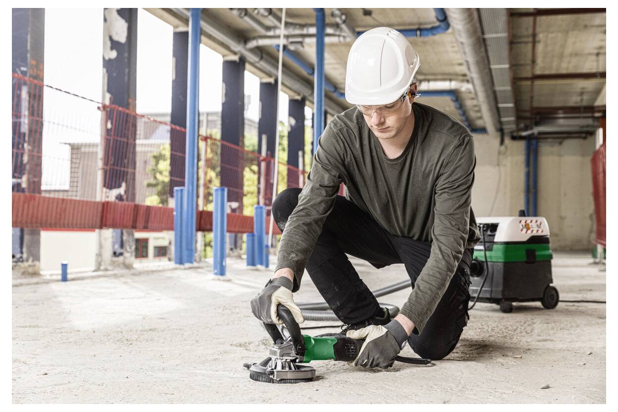 Bauarbeiter mit Schutzhelm hockt sich hin und bedient ein tragbares Elektrowerkzeug auf einem Betonboden auf einer Indoor-Baustelle.