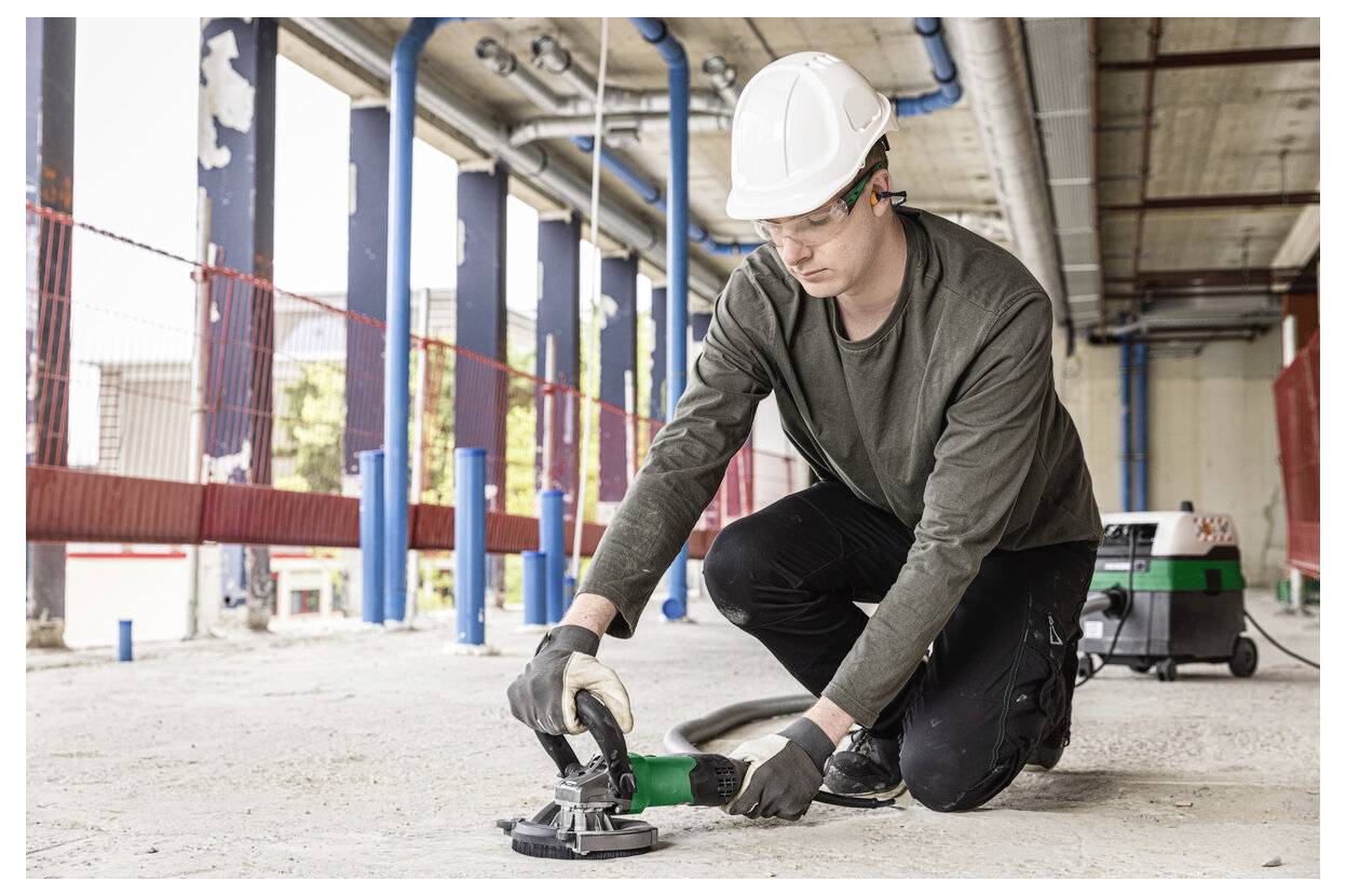 Arbeiter auf einer Baustelle kniet in Arbeitskleidung und nutzt ein Elektrowerkzeug, um Beton zu schneiden oder zu schleifen.