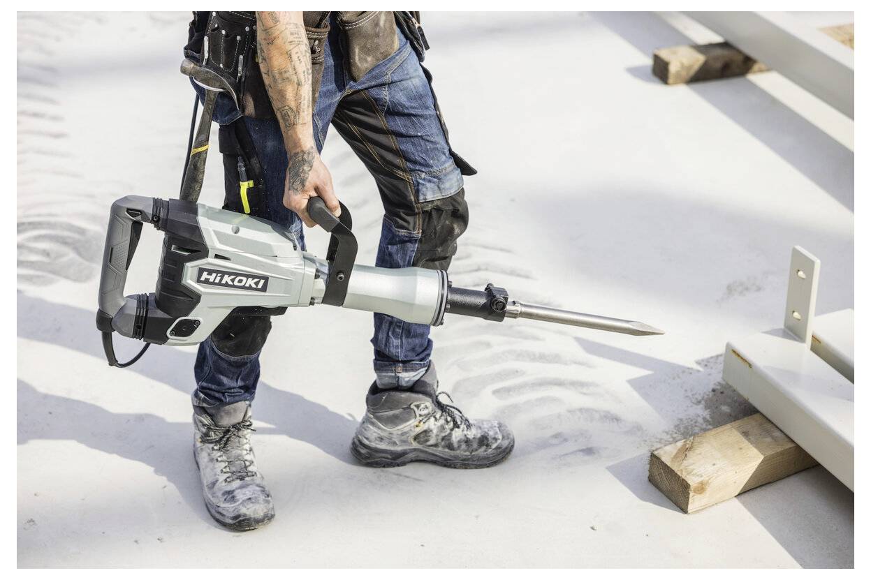 Arbeiter, der einen Hikoki-Abbruchhammer verwendet, um Beton neben einem Metallrahmen zu zerbrechen, mit Staub und Blöcken auf dem Boden.