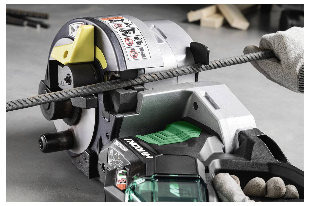 Eine Person benutzt eine Makita-Bewehrungsschneidemaschine, um einen Stahlstab zu schneiden, wobei handschuhtragene Hände das Werkzeug und den Stab auf einem Werkstattboden halten.