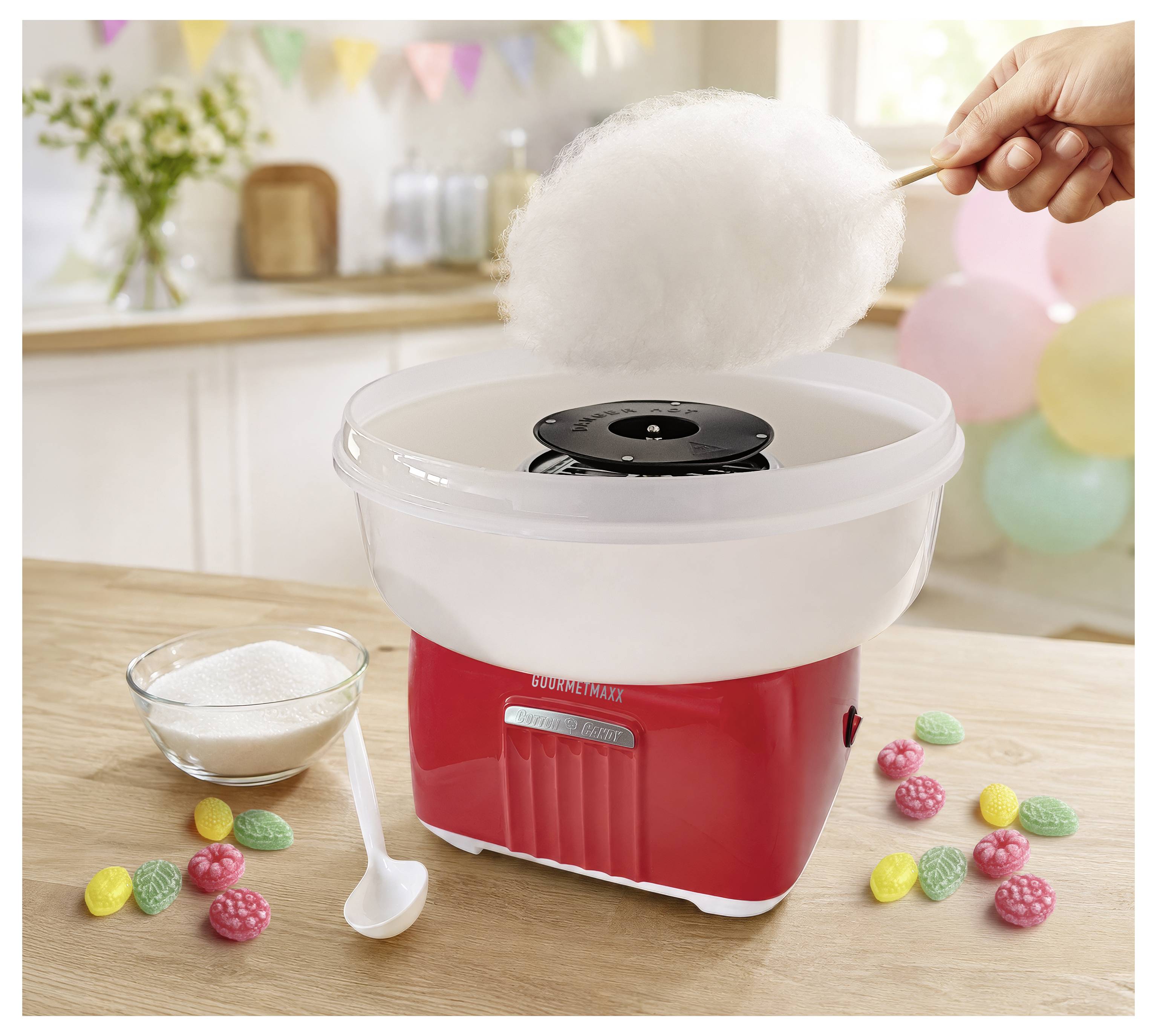 Eine Zuckerwattemaschine spinnt flauschige weiße Zuckerwatte auf einen Stab, mit einer Zuckerschale, einem Löffel und bunten Bonbons auf einem Tisch.