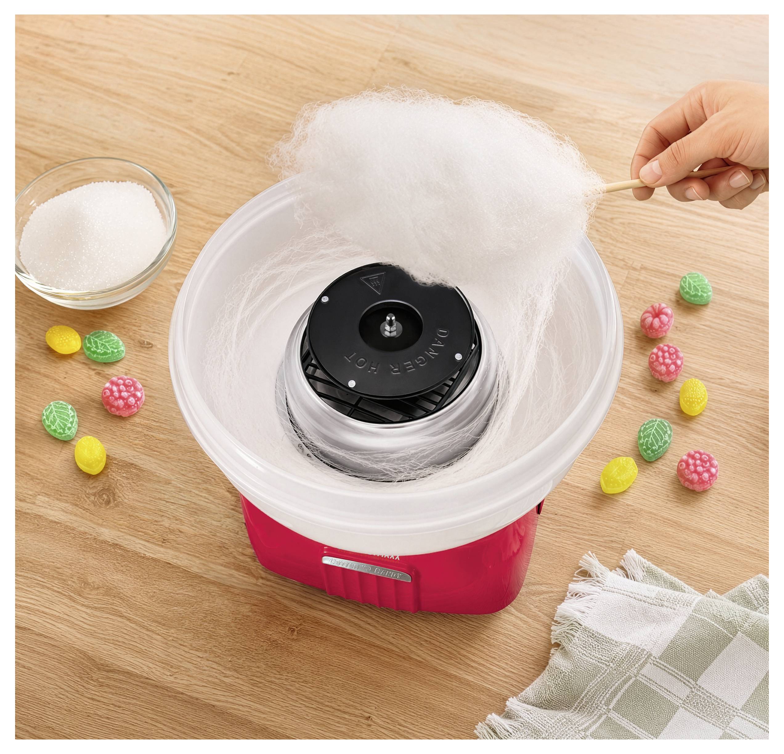 Zuckerwattemaschine, die weiße Zuckerwatte spinnt, während eine Hand Zucker hinzufügt; Schüssel und bunte Bonbons auf einem Holztisch.