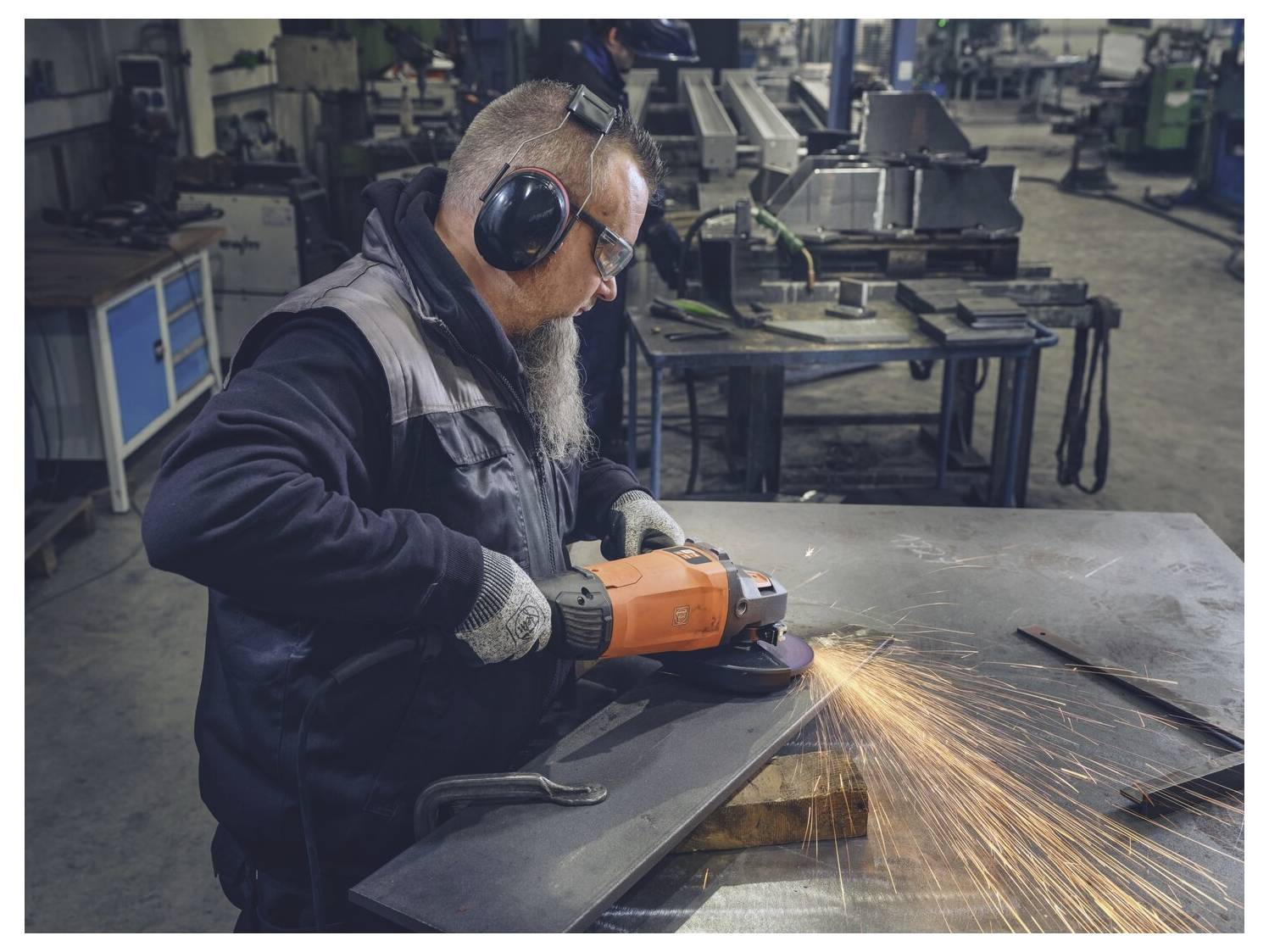 Arbeiter mit Gehörschutz schleifen ein Metallblech mit einem Winkelschleifer und senden Funken über eine Industriewerkstatt.