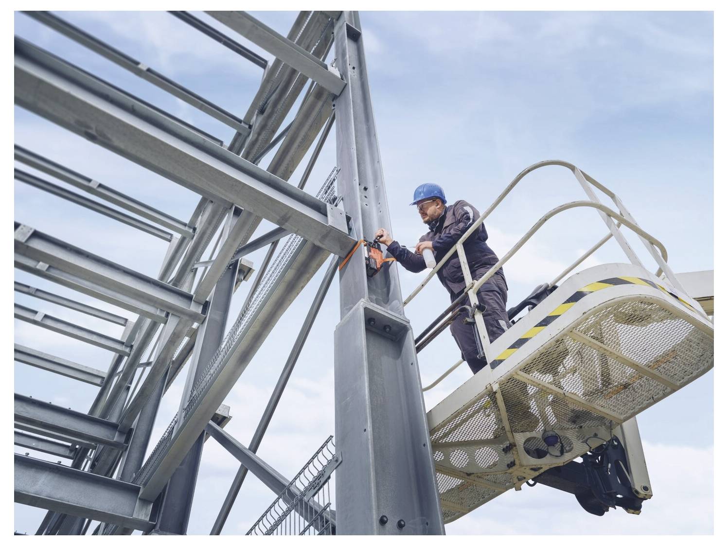 Ein Arbeiter in einem blauen Schutzhelm sichert Stahlträger von einer erhöhten Plattform auf einer Baustelle.
