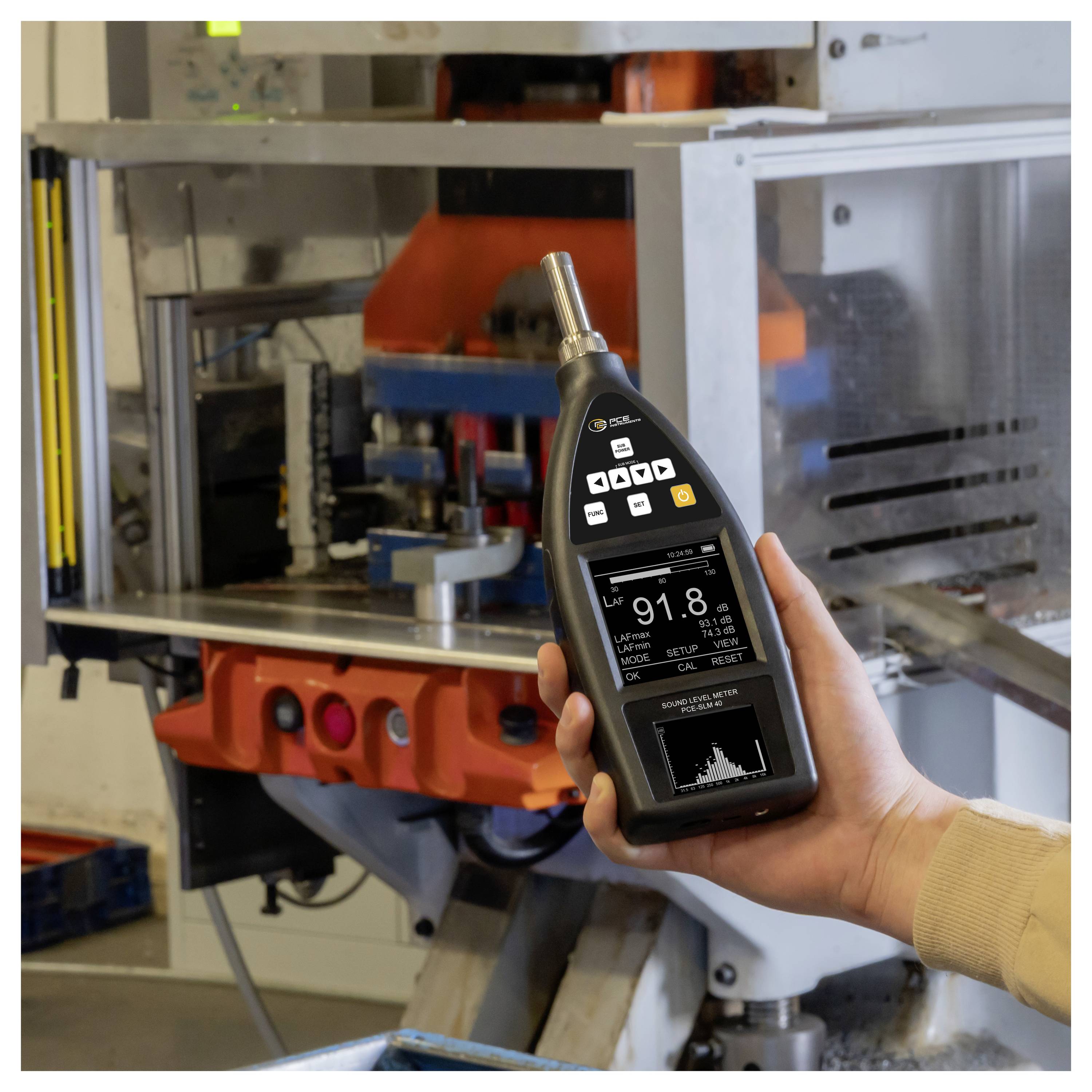 Hand, die ein tragbares Messinstrument vor Industriemaschinen hält, das Messwerte und ein Diagramm auf seinem Bildschirm anzeigt.