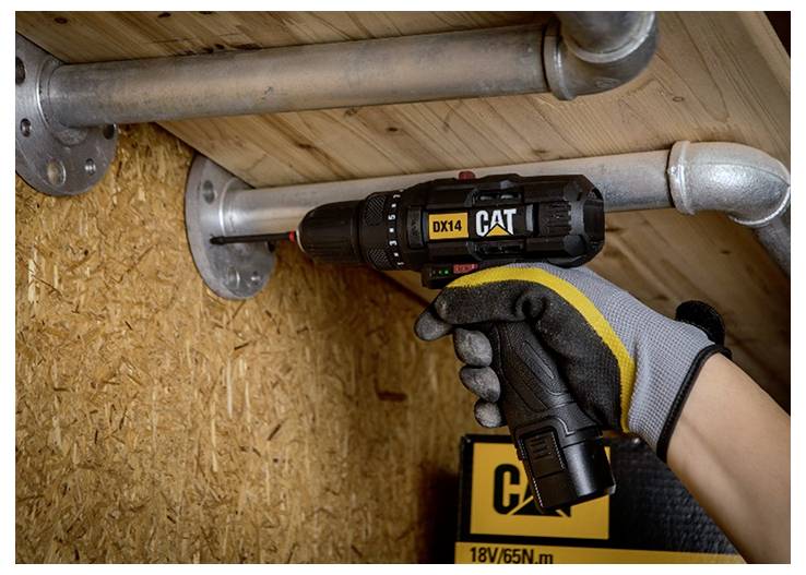 Behandschuhte Hand benutzt einen CAT-Akkubohrer, um eine Metallrohrverbindung in einem Holzrahmenwerk zu befestigen, wobei ein Werkzeugetikett mit 18V/65N·m sichtbar ist.