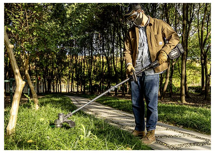 Ein Mann benutzt einen kabellosen Fadentrimmer, um Gras entlang eines Parkweges zu schneiden, der von Bäumen umgeben ist.