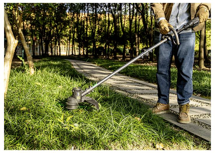 Eine Person nutzt einen Rasentrimmer, um Gras entlang eines Gehwegs im Park an einem sonnigen Tag zu schneiden.