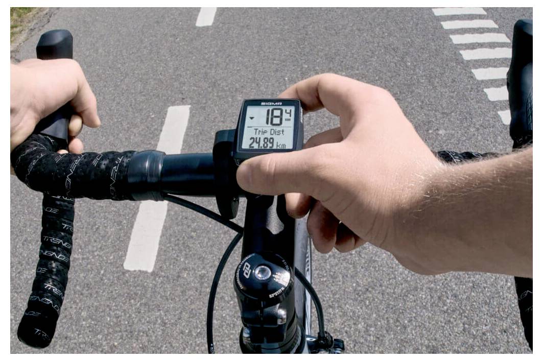 Die Hände eines Radfahrers liegen auf dem Lenker und stellen einen Fahrradcomputer ein, der 18,4 und eine Fahrstrecke von 24,89 km anzeigt, während er auf einer Straße fährt.