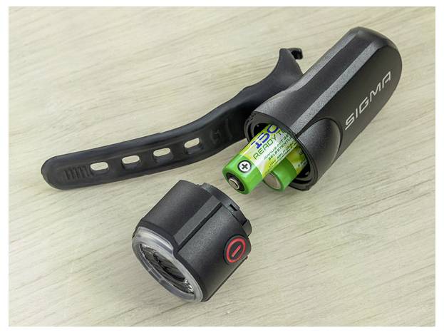 Zerlegter Sigma-Fahrradscheinwerfer mit entfernten Batterien, das Lampengehäuse, die Objektivkappe und den schwarzen Befestigungsriemen auf einer hellen Holzoberfläche zeigend.