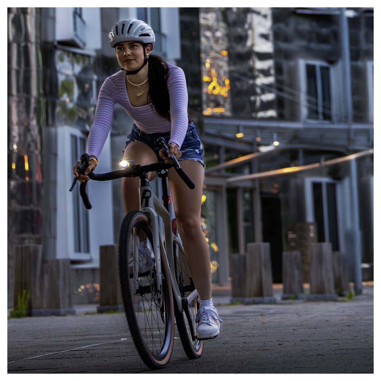 Junge Frau fährt mit dem Rennrad auf einem städtischen Gehweg in der Dämmerung; sie trägt Helm, gestreiftes Oberteil und Shorts.