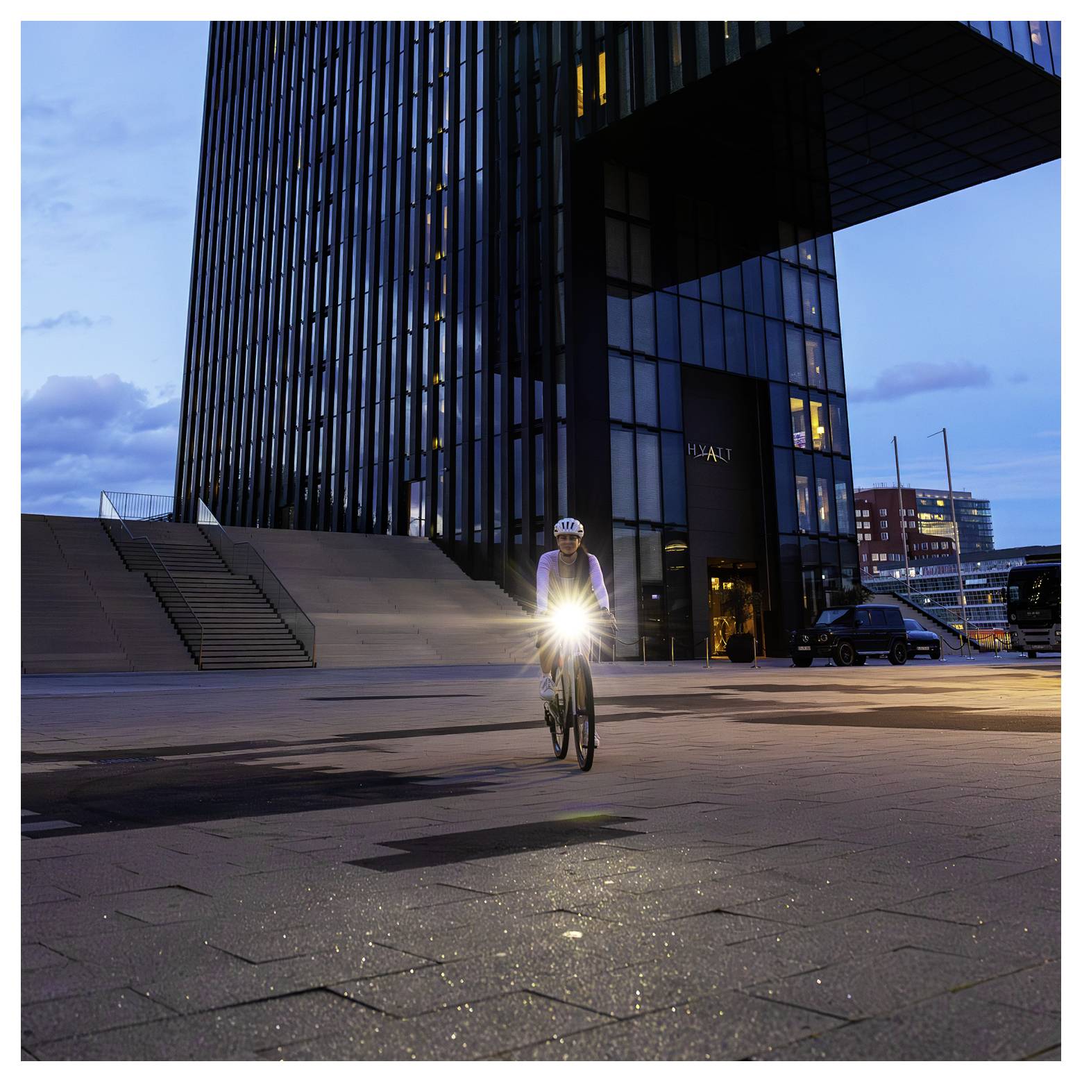 Radfahrer mit eingeschaltetem Frontlicht fährt vor einem modernen Hochhaus bei Dämmerung auf einem leeren Platz.