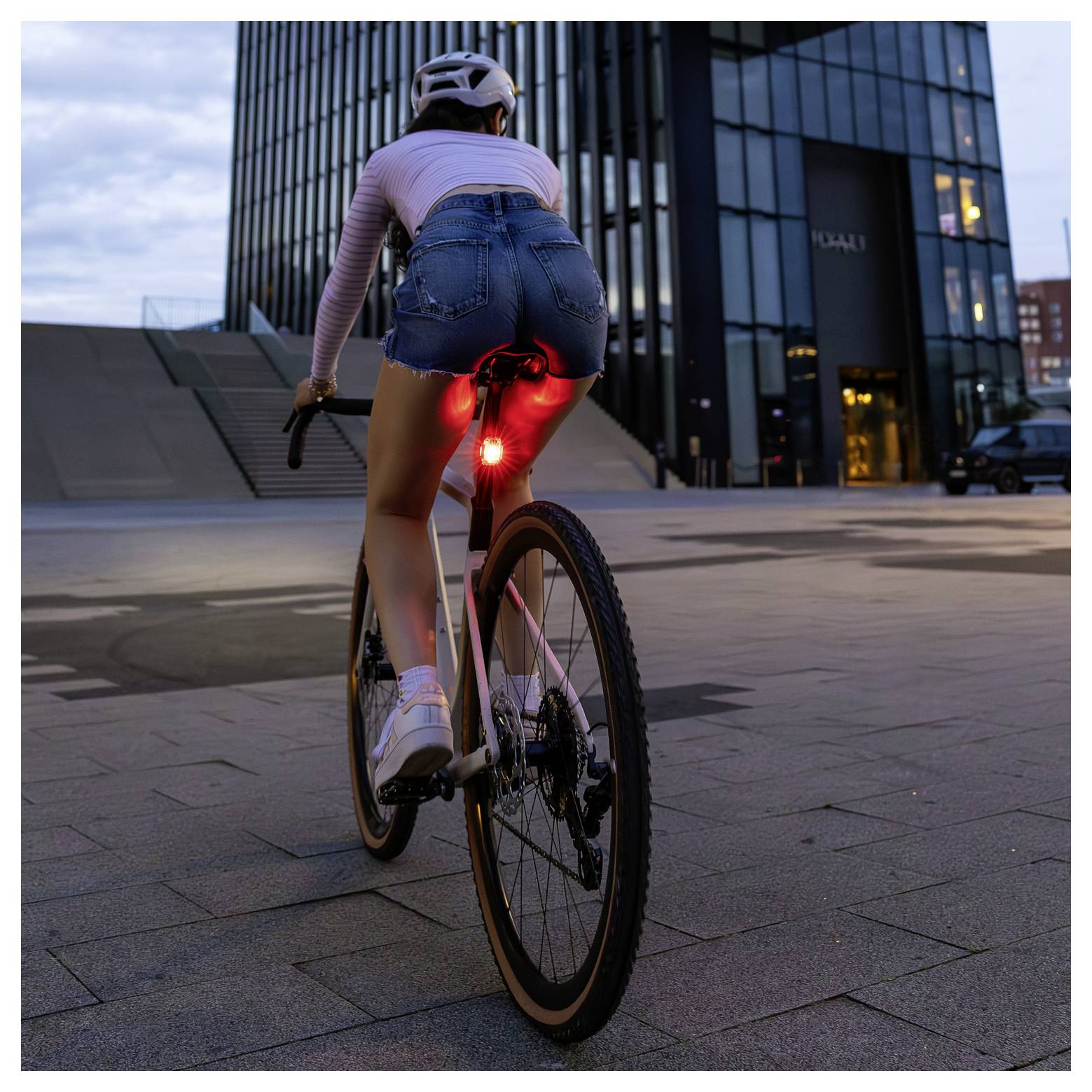 Radfahrerin fährt bei Dämmerung vor modernen Bürogebäuden; rotes Rücklicht am Fahrrad leuchtet deutlich.