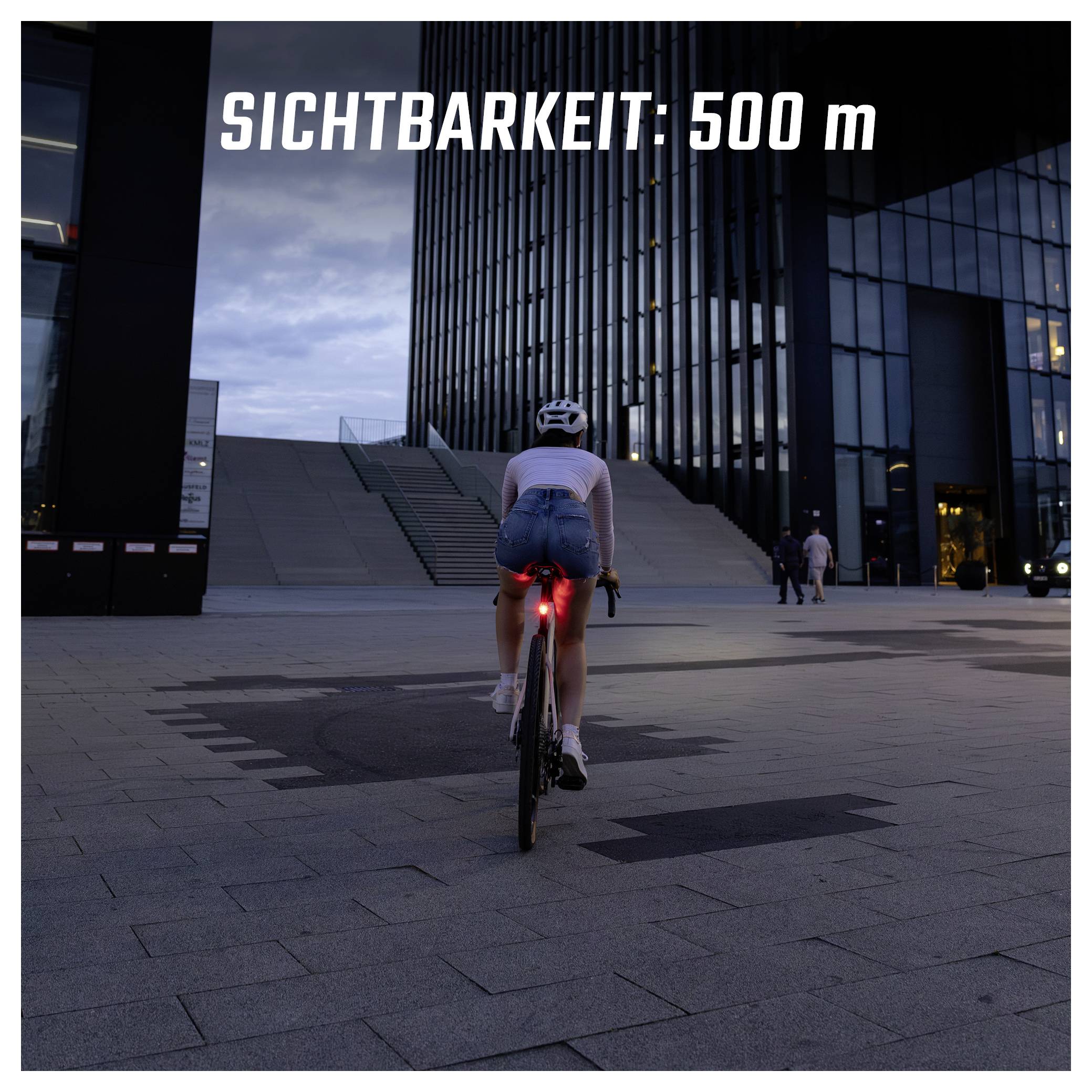 Radfahrerin vor moderner Gebäudefassade in der Dämmerung, mit rotem Rücklicht; Text im Bild: 'SICHTBARKEIT: 500 m'.