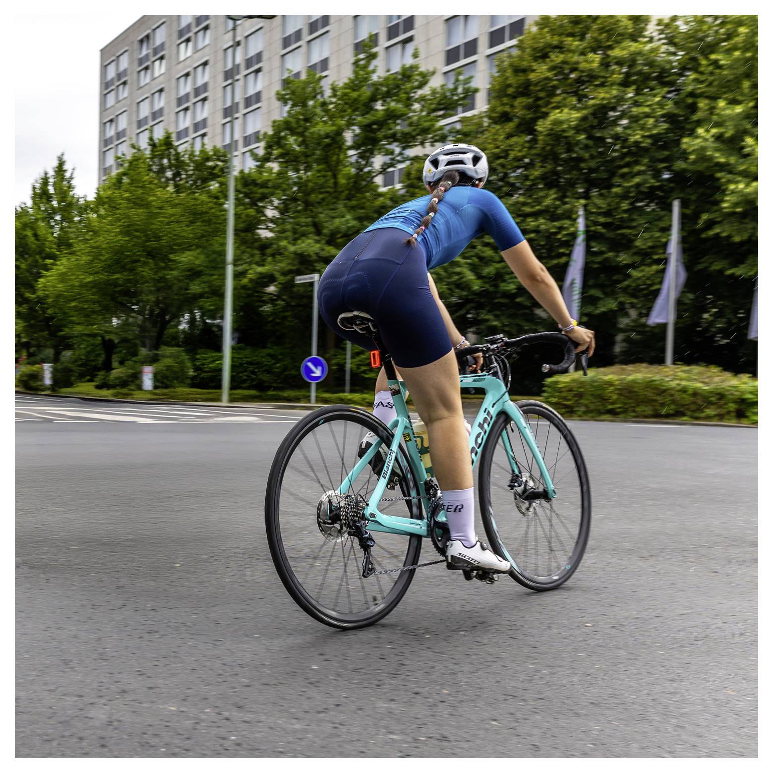 Ein Radfahrer in Blau, der ein türkisfarbenes Rennrad auf einer Stadtstraße fährt, von hinten fotografiert in der Nähe von Bäumen und Wohngebäuden.
