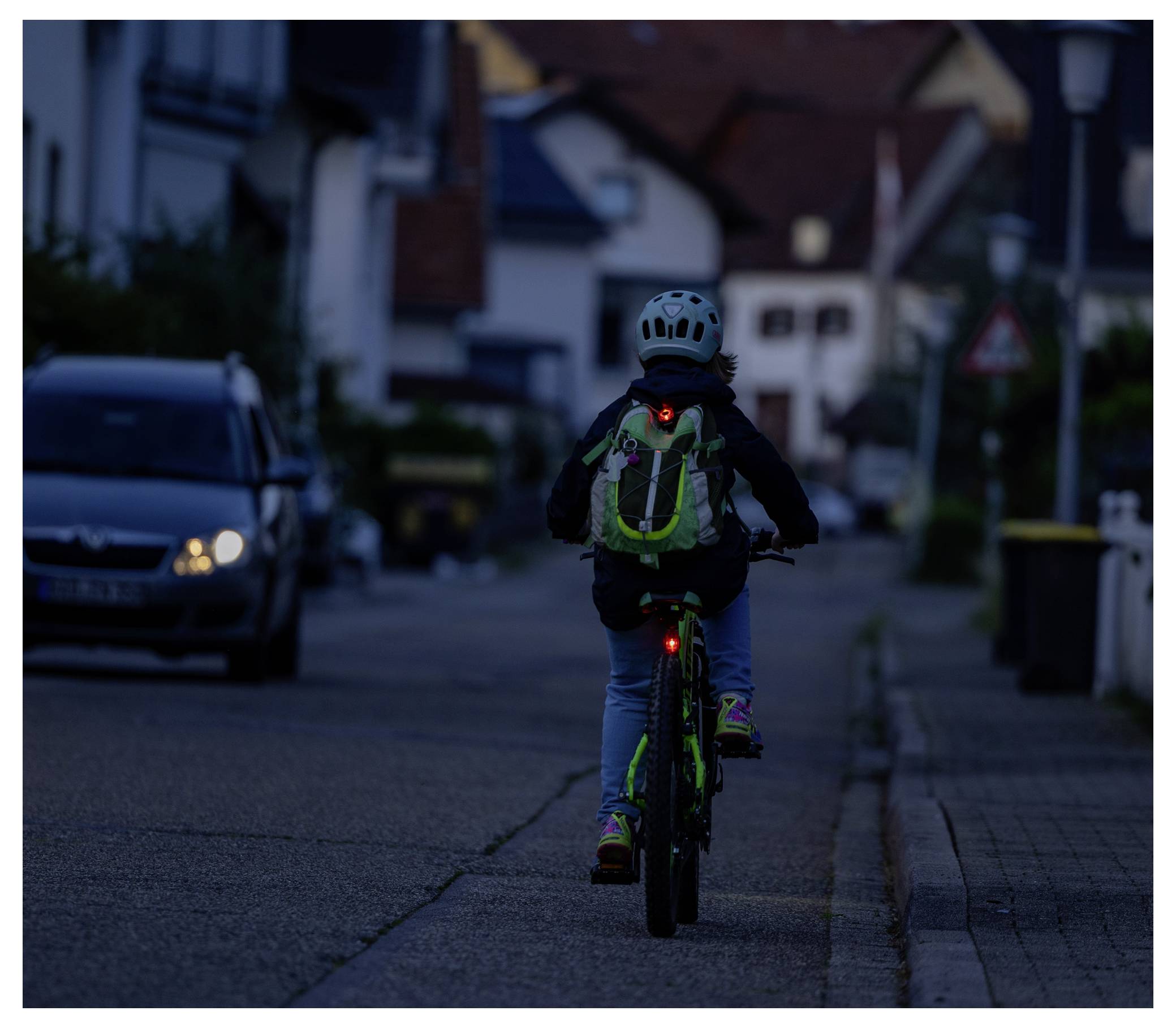 Ein Kind fährt auf einem Fahrrad eine schwach beleuchtete Wohnstraße in der Dämmerung hinunter, trägt einen Helm und einen Rucksack mit einem sichtbaren roten Rücklicht.