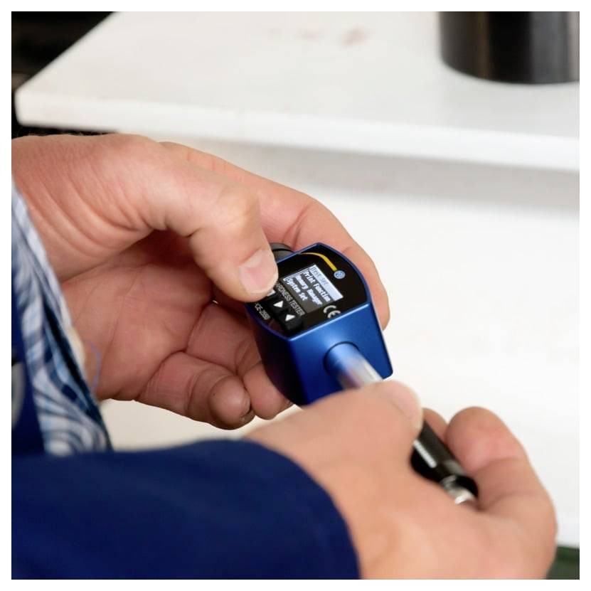 Hände, die ein kleines blaues digitales Messinstrument halten, während eine Sonde darin auf einer weißen Arbeitsfläche eingeführt wird.
