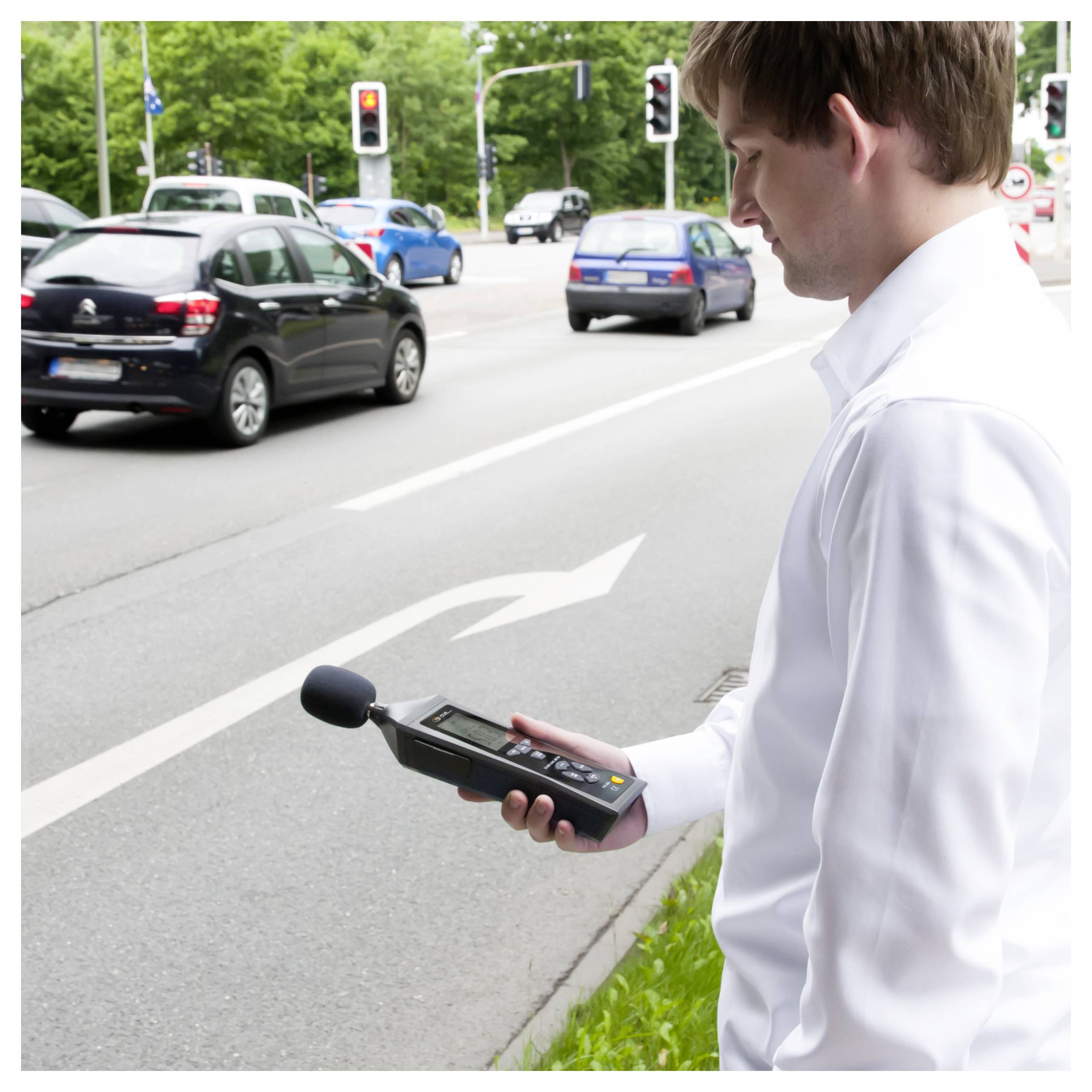 Ein Mann, der neben einer belebten Straße steht, nutzt ein tragbares Schallpegelmessgerät, um den Verkehrslärm in der Nähe einer Kreuzung zu messen.