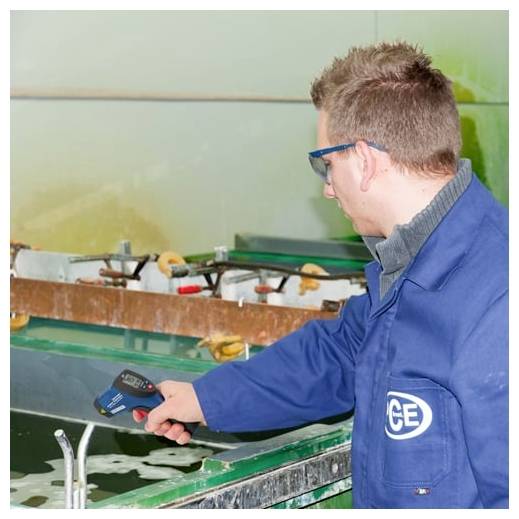 Techniker im blauen Laborkittel benutzt ein tragbares Testgerät über einem Wasserbehälter in einer industriellen oder Laborumgebung.