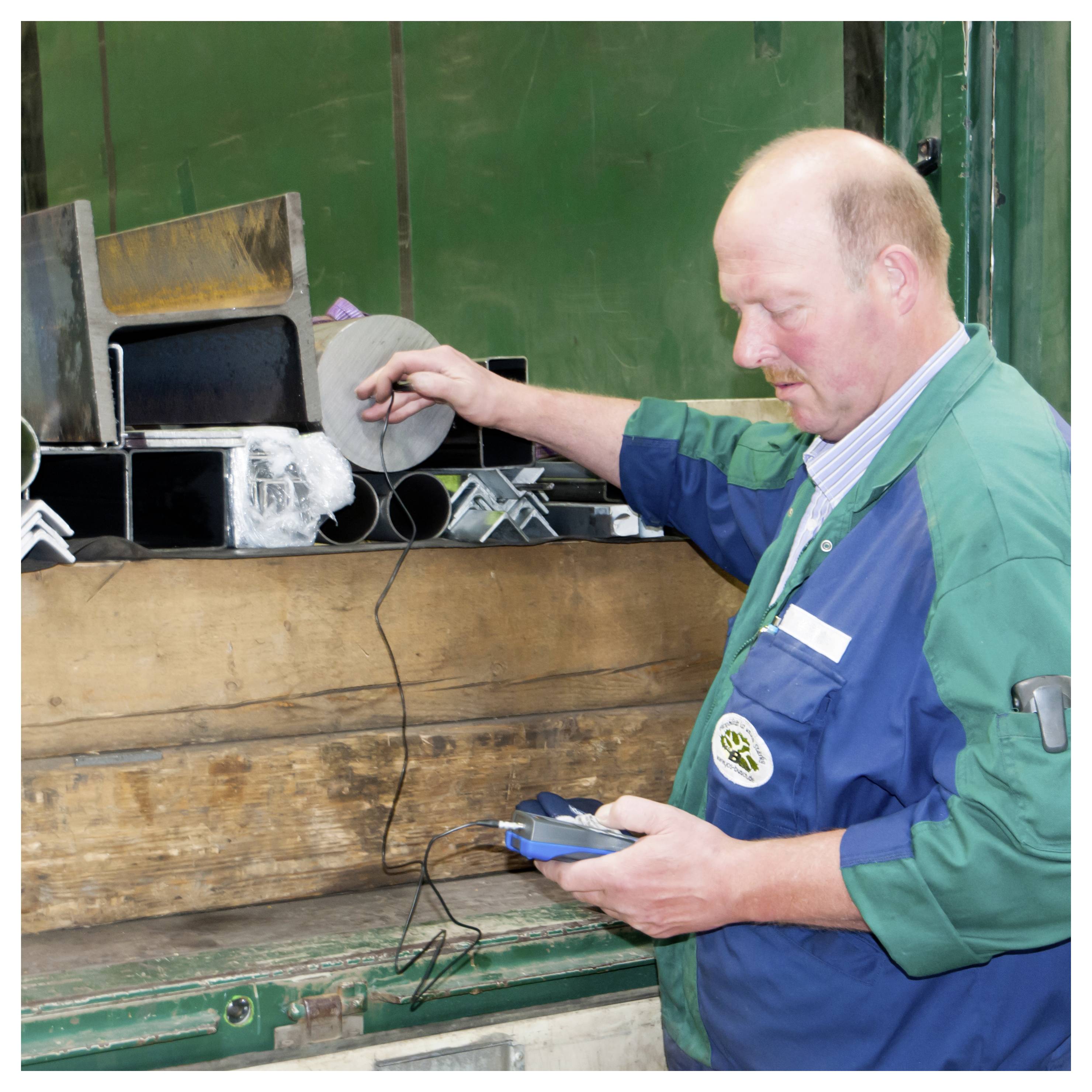 Arbeiter in einer Werkstatt inspiziert Metallteile mit einem tragbaren Gerät neben einer Werkbank voller Werkzeuge und Materialien.