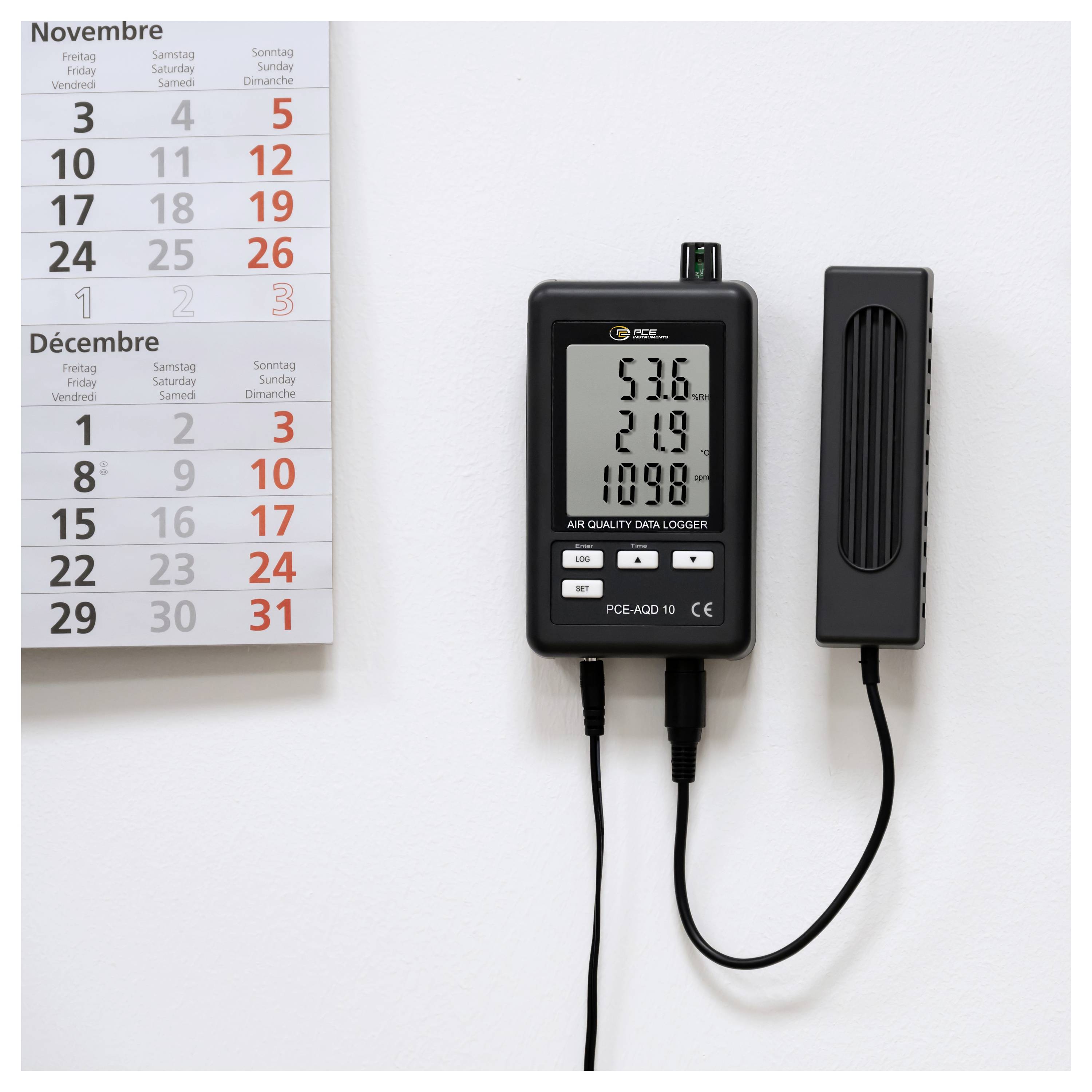 Wandmontiertes Luftqualitätsdaten-Erfassungsgerät mit digitaler Anzeige, die 53,6, 21,9 und 1098 zeigt, neben einer hängenden Kalenderseite für November und Dezember.
