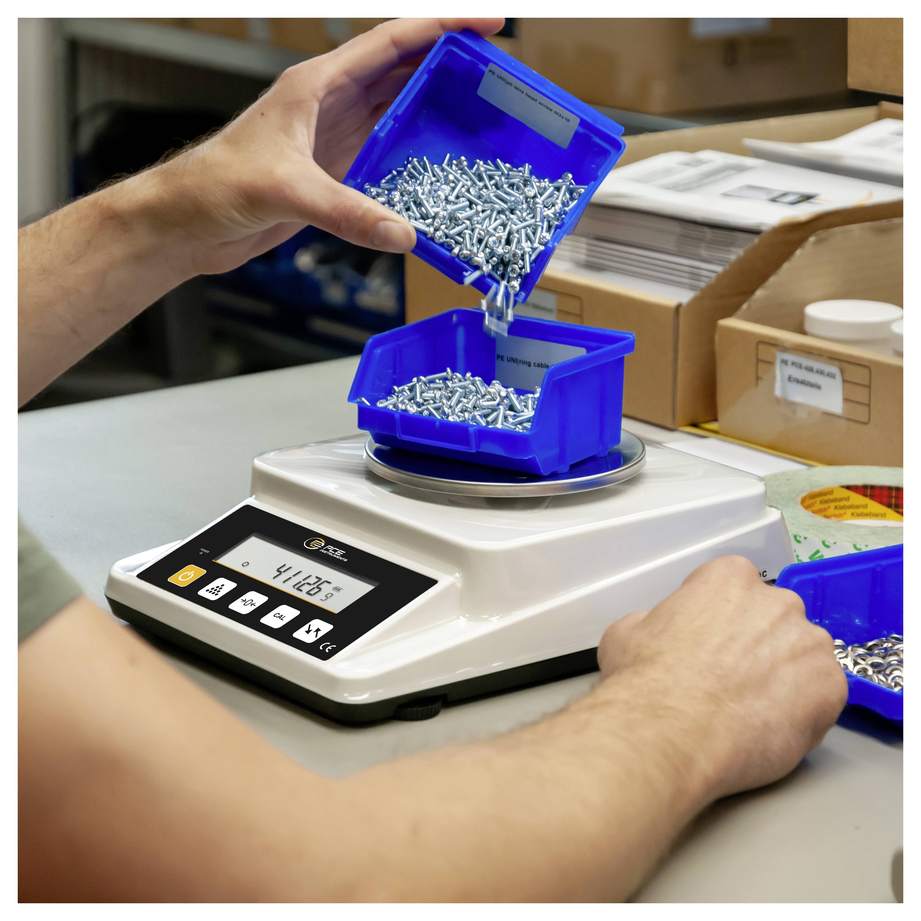 Eine Person schüttet kleine Metallschrauben aus einem blauen Behälter auf eine digitale Waage in einer Werkstatt oder einem Lagerverwaltungs-Sortiersystem.