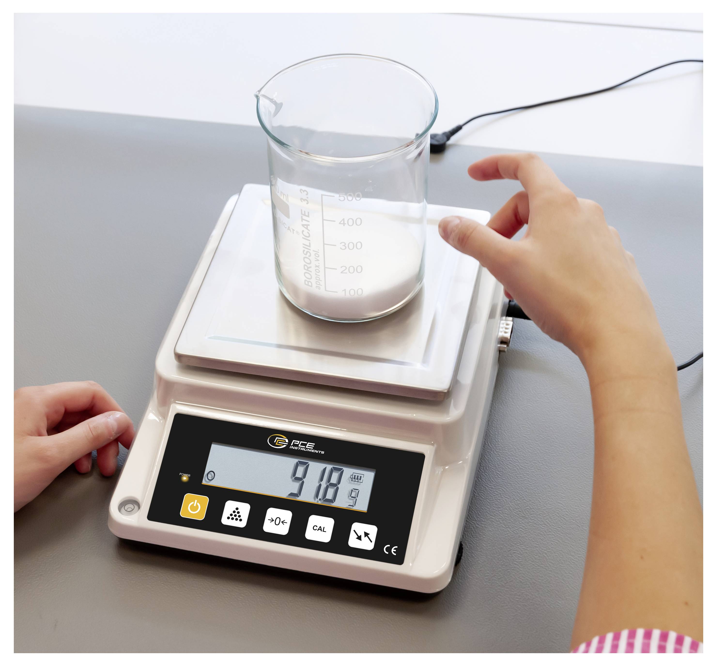 Ein Becher mit weißem Pulver sitzt auf einer digitalen Laborwaage, während eine Person diese justiert; die Anzeige zeigt 918 g.
