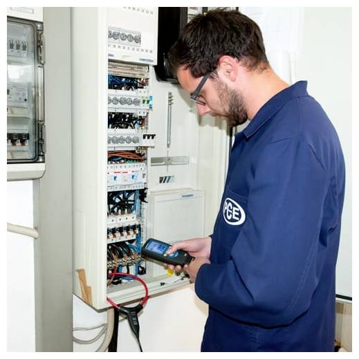 Techniker in blauer Uniform inspiziert ein offenes Elektrosteuerschrank mit einem tragbaren Prüfgerät.