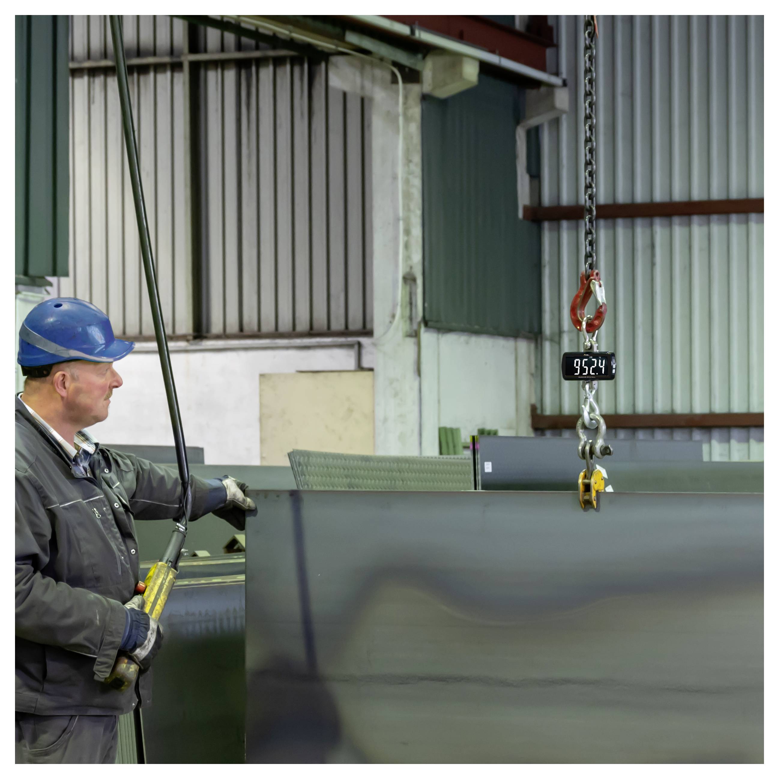 Fabrikarbeiter in einem Schutzhelm, der ein großes Stahlblech leitet, das von einem Laufkran in einer Industriewerkstatt angehoben wird.