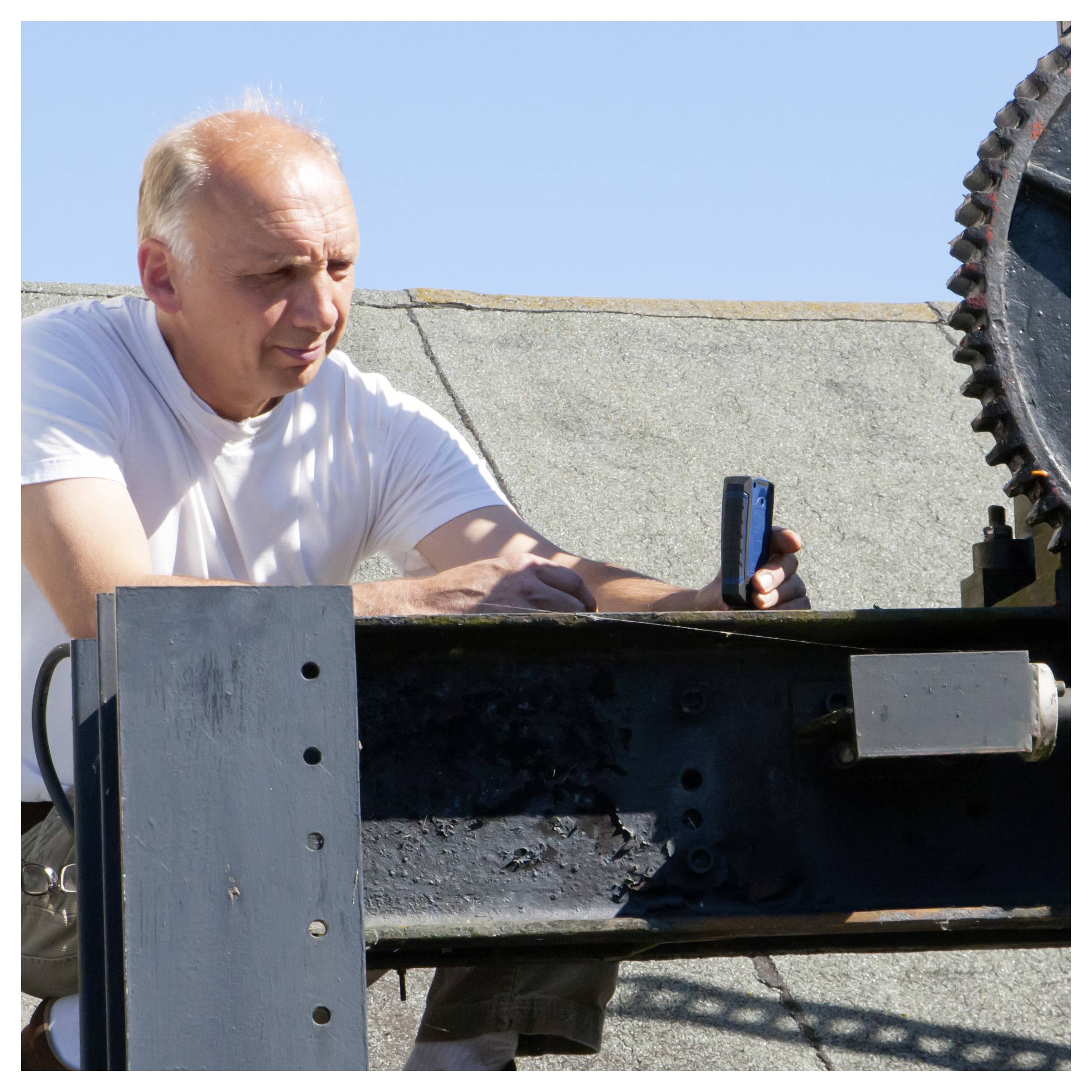 Ein älterer Mann in einem weißen T-Shirt arbeitet im Freien mit einer großen Metallsäge oder Schneidemaschine, hält ein Werkzeug und inspiziert die Einrichtung.