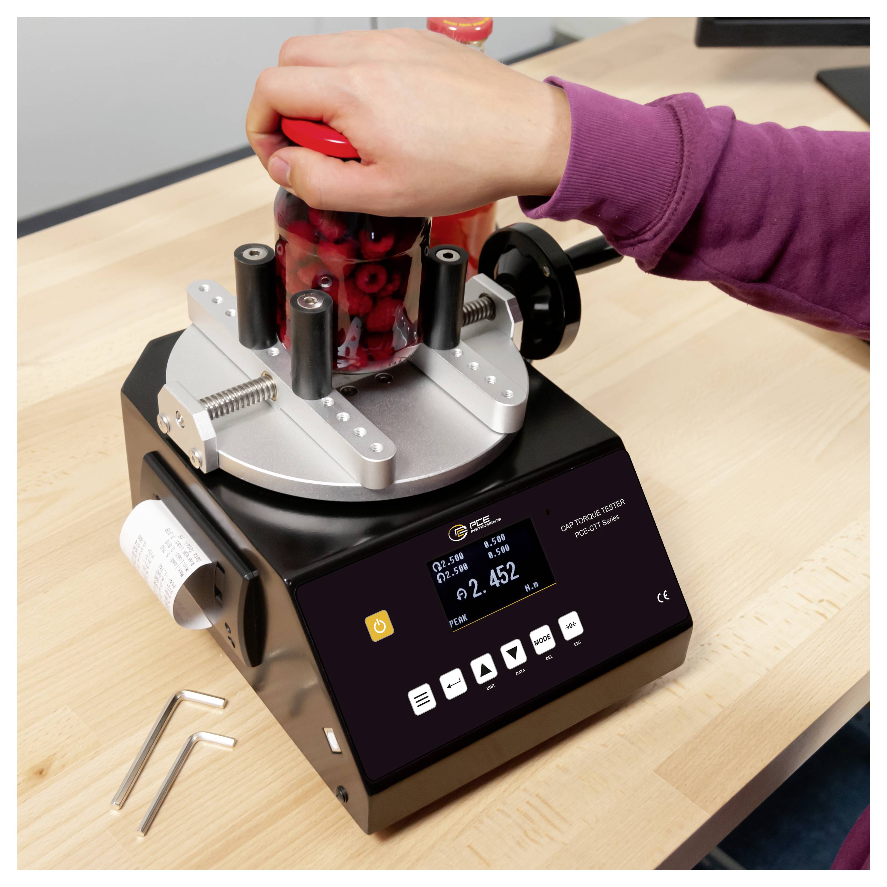 Person dreht kurbelt an einem roten Glas mit digitaler Verschlussdeckel-Drehmomentprüfer auf einer Workbench, mit Innensechskantschlüsseln in der Nähe.