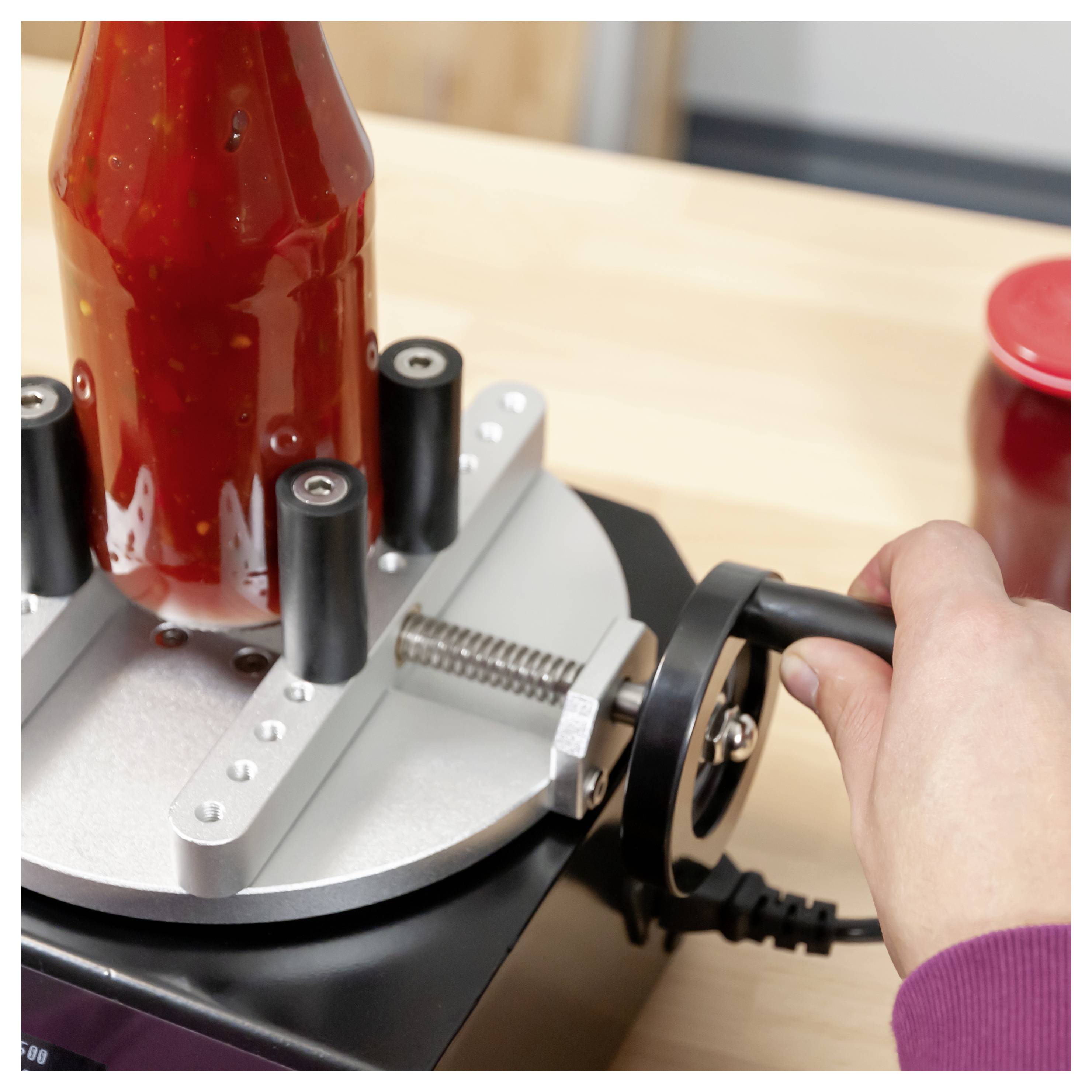 Eine Hand dreht die Kurbel einer Flaschenettiquettiermaschine und wickelt ein Etikett um eine rote Flasche auf einer Werkbank.