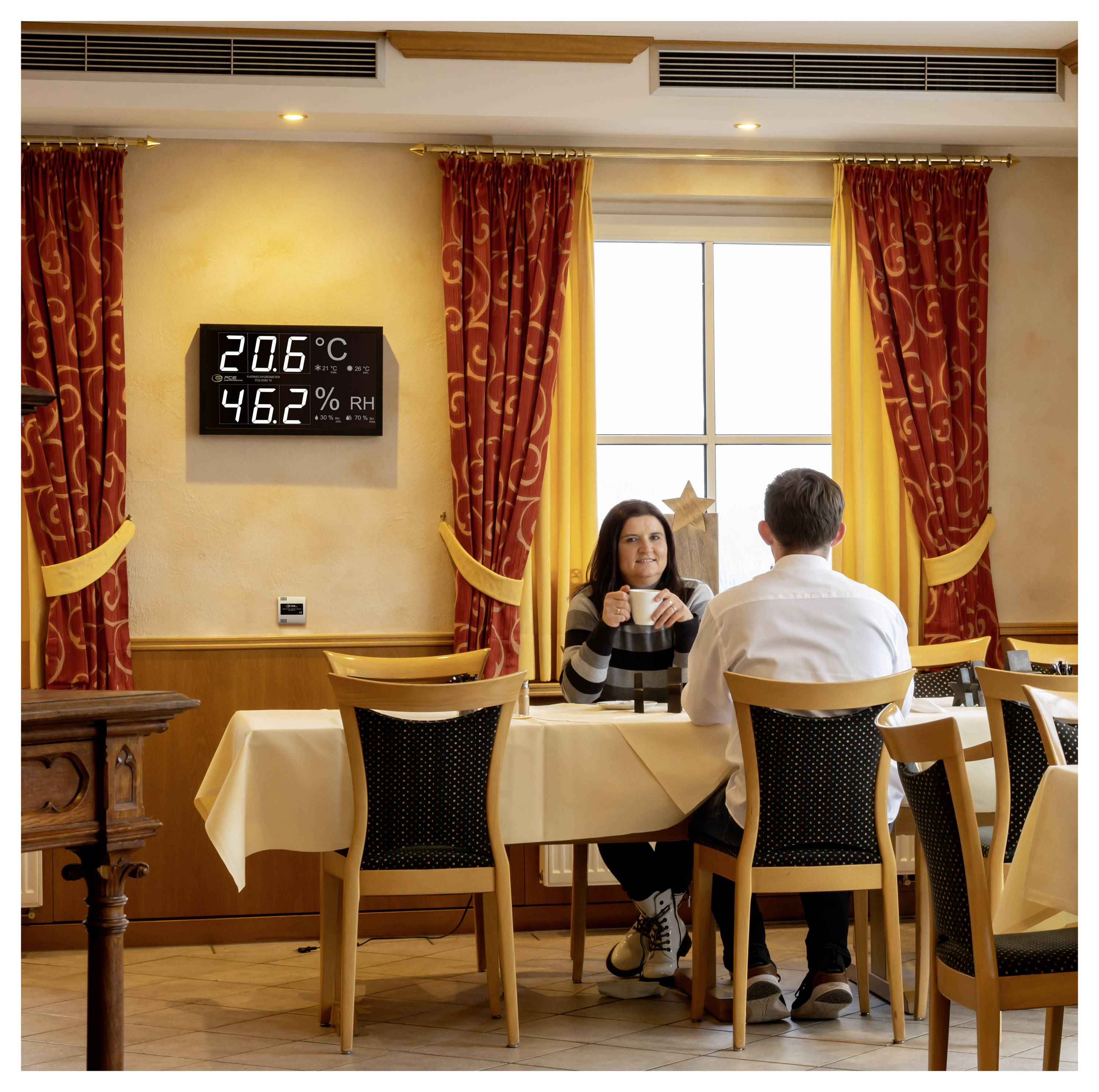 Zwei Personen sitzen sich an einem Restauranttisch neben einem Fenster gegenüber; eine Wandanzeige zeigt „20,6°C