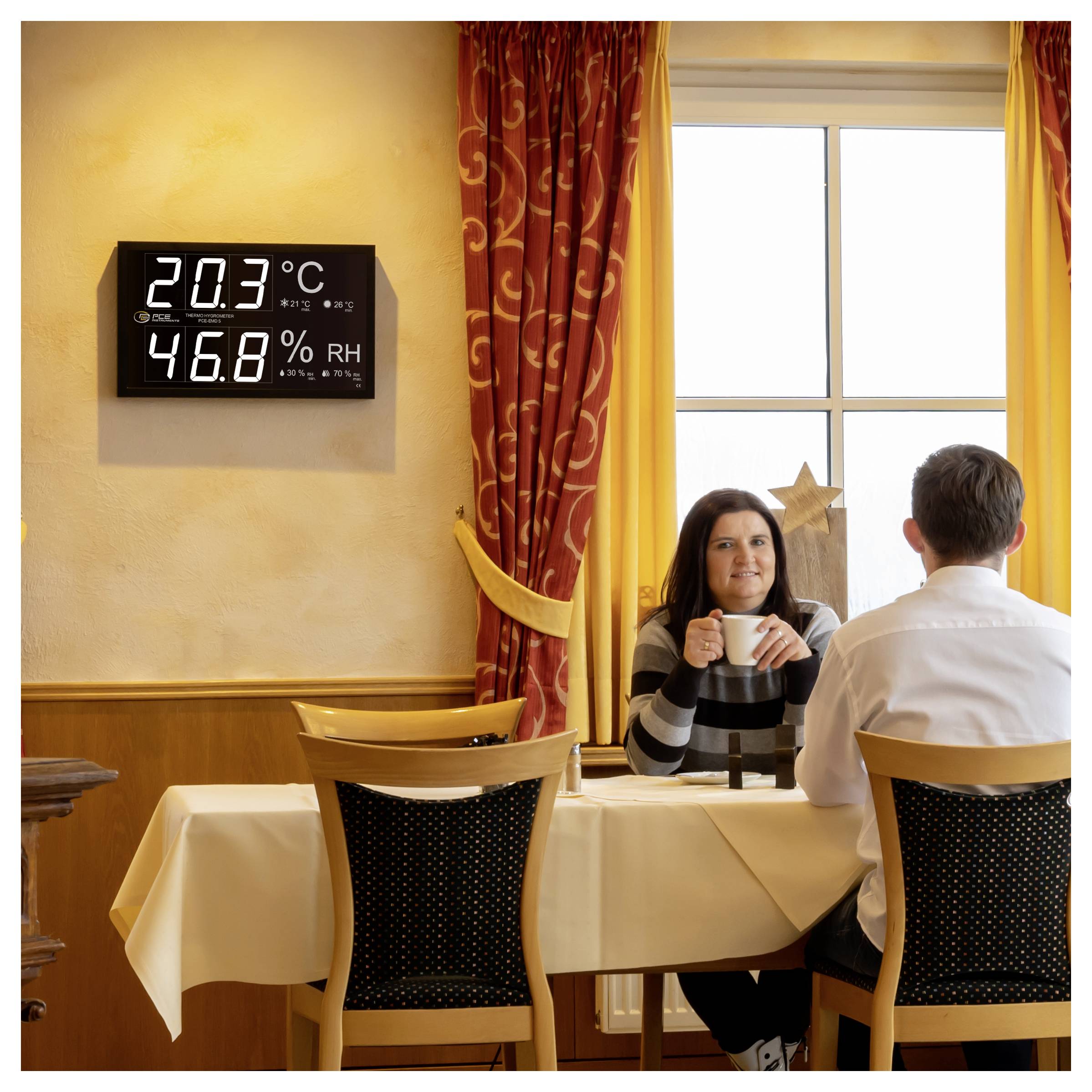 Eine Frau und ein Mann sitzen an einem Tisch in einem Restaurant neben einem Fenster, während eine Wandanzeige 20,3°C und 46,8% relative Luftfeuchte anzeigt.