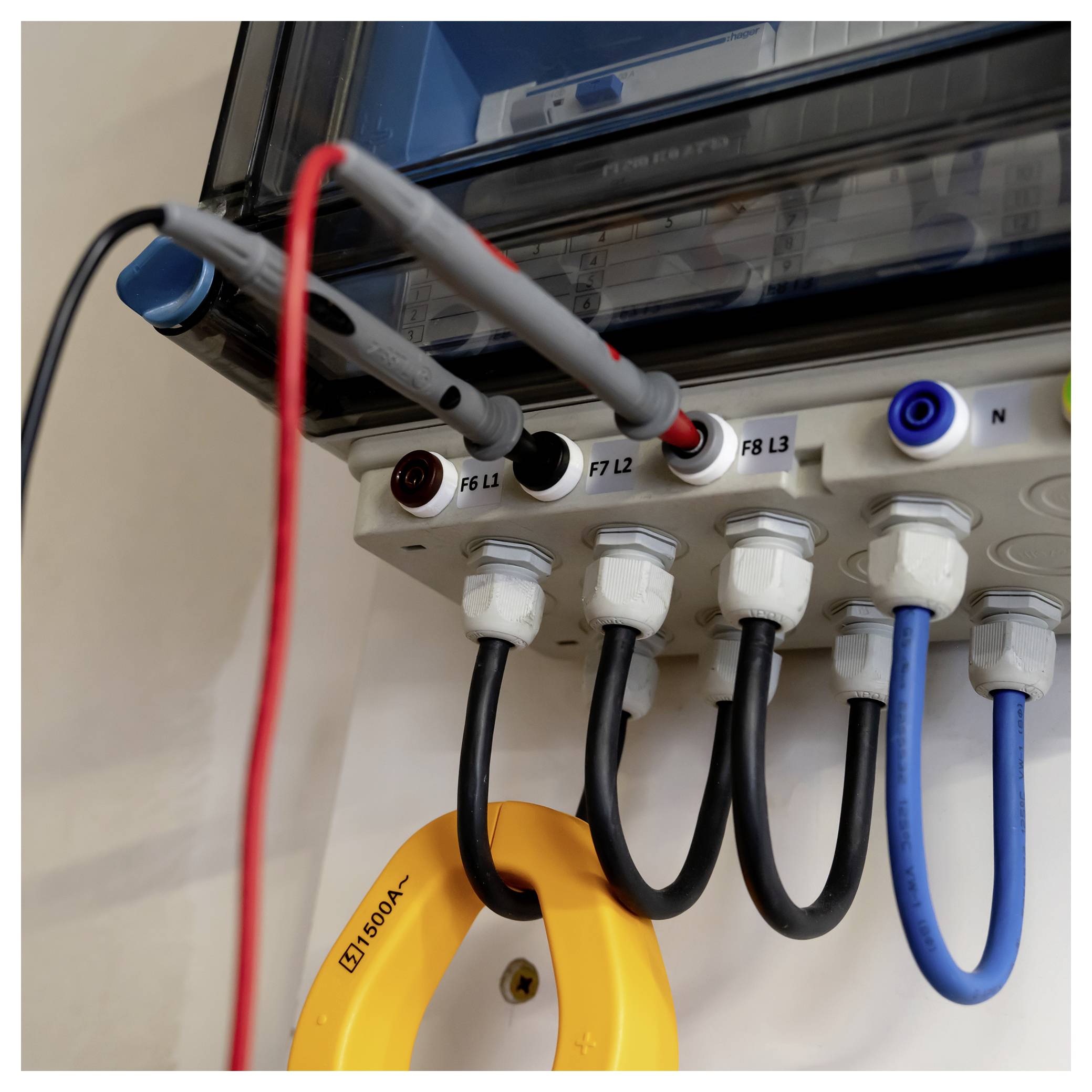Nahaufnahme eines elektrischen Verteilerpanels mit angeschlossenen Stromkabeln, beschrifteten Anschlüssen und einer gelben Zangenmesszange um einen Draht.
