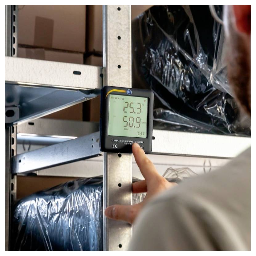 Eine Person überprüft ein digitales Thermometer auf einem Metallregal; das Display zeigt 25.3 und 50.9, wahrscheinlich Temperatur und Luftfeuchtigkeit.