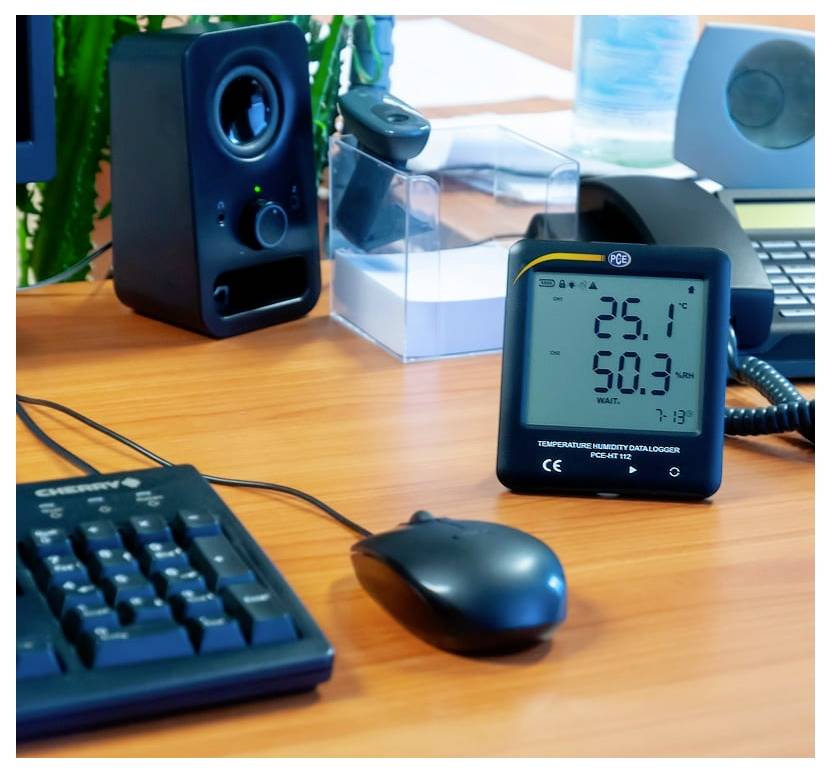 Ein digitales Temperatur- und Feuchtigkeitsmessgerät auf einem Schreibtisch zeigt 25,1°C und 50,3% Luftfeuchtigkeit an, neben einer Tastatur, einer Maus und einem Lautsprecher.