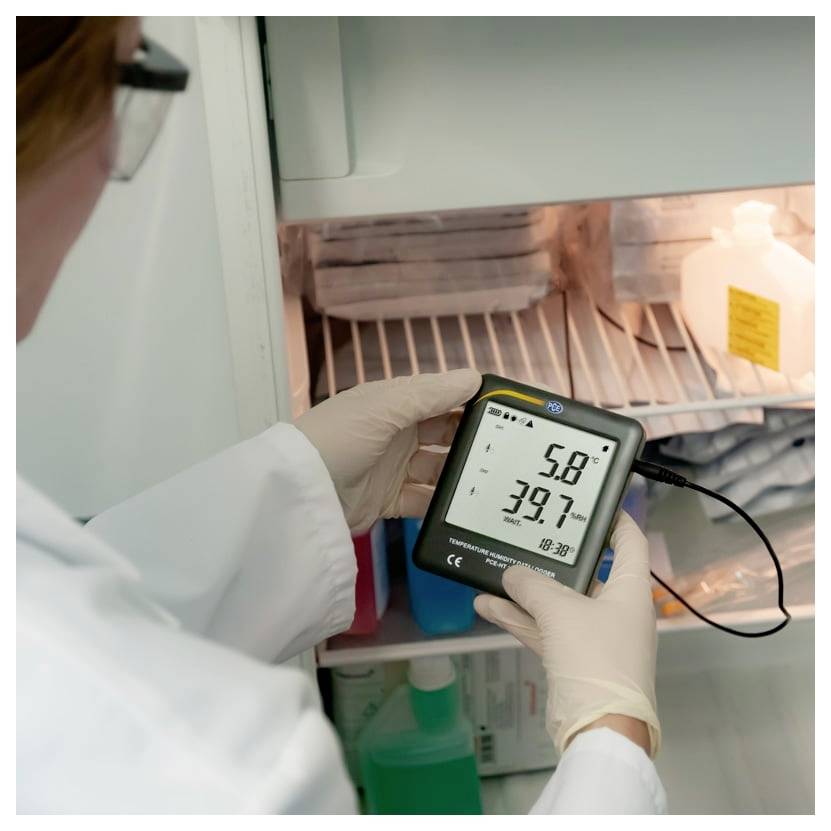 Laborant mit Handschuhen überprüft ein digitales Thermometer-Hygrometer in einem Kühlschrank, das 5,8 und 39,7 anzeigt, mit einer angeschlossenen Sonde.