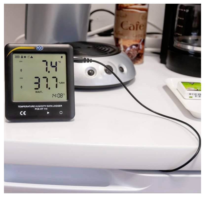 Digitales Temperatur- und Feuchtigkeitsmessgerät auf einem Schreibtisch zeigt 7,4°C und 37,7% rel. Feuchte an, mit einem Kabel verbunden mit nahegelegener Ausrüstung.
