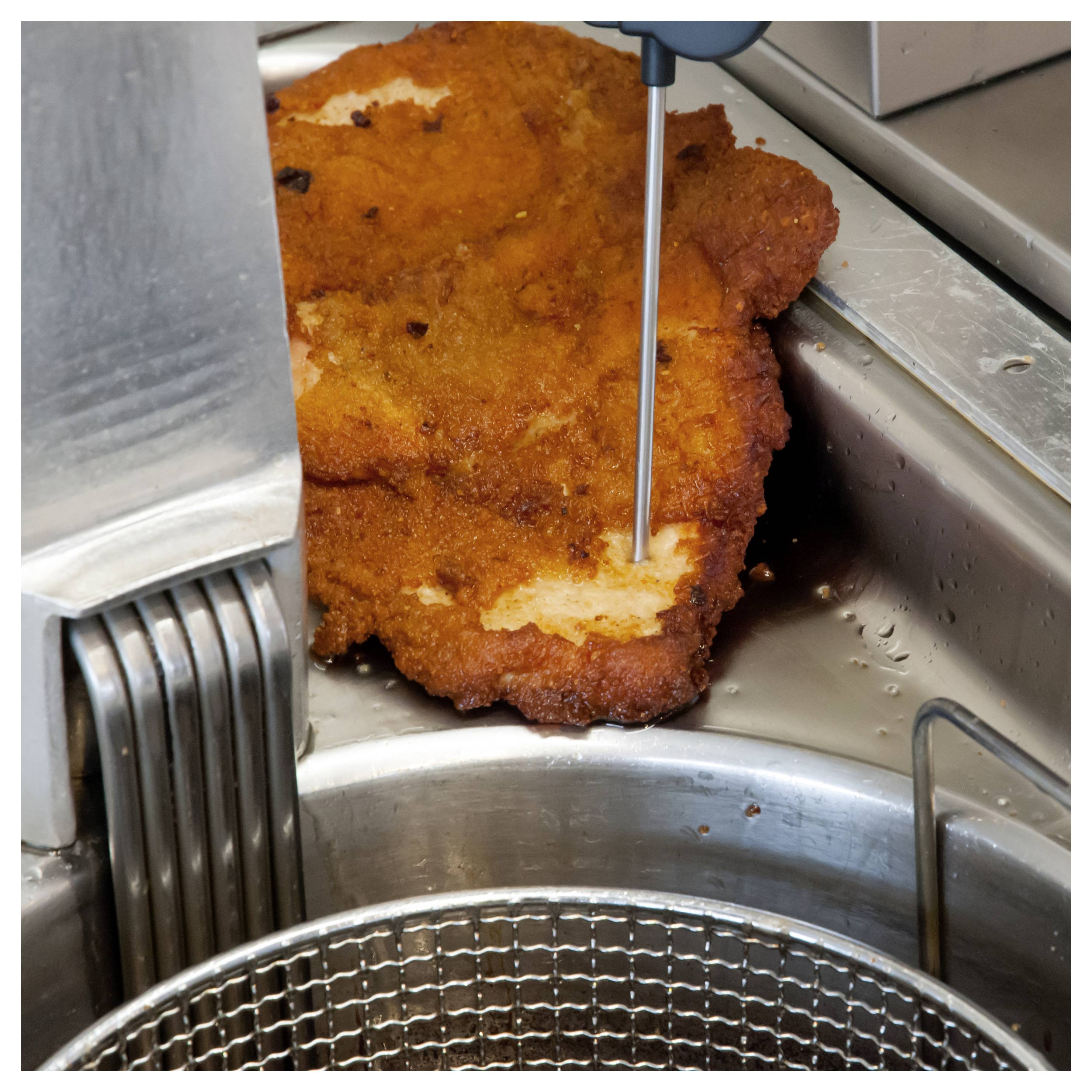 Ein paniertes Schnitzel wird von einer Metallthermometer-Sonde in der Nähe einer Fritteuse durchbohrt, um seine Gartemperatur zu überprüfen.
