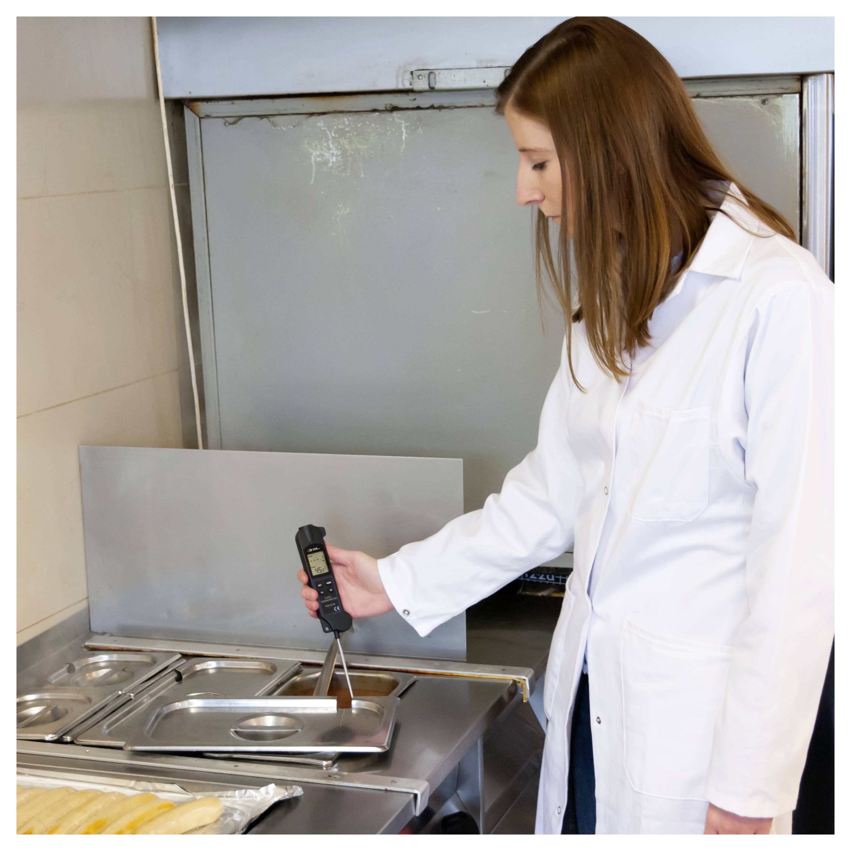 Ein Techniker im Laborkittel misst die Temperatur mit einer digitalen Sonde über einem Edelstahllebensmitteltablett in einer Küche oder einem Lebensmitteltestbereich.