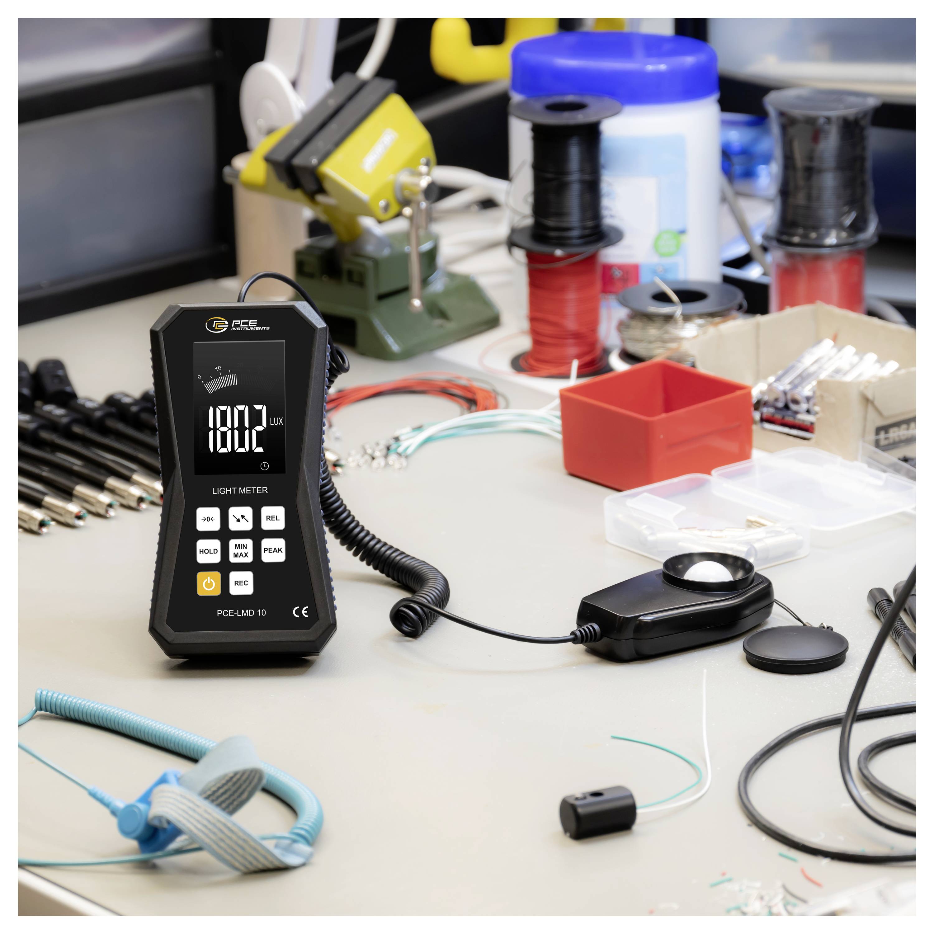 Digitales Luxmeter zeigt 1802 Lux auf einer Werkbank neben Spulen, Werkzeugen, Drähten und kleinen elektronischen Komponenten in einem Labor oder einer Werkstatt an.