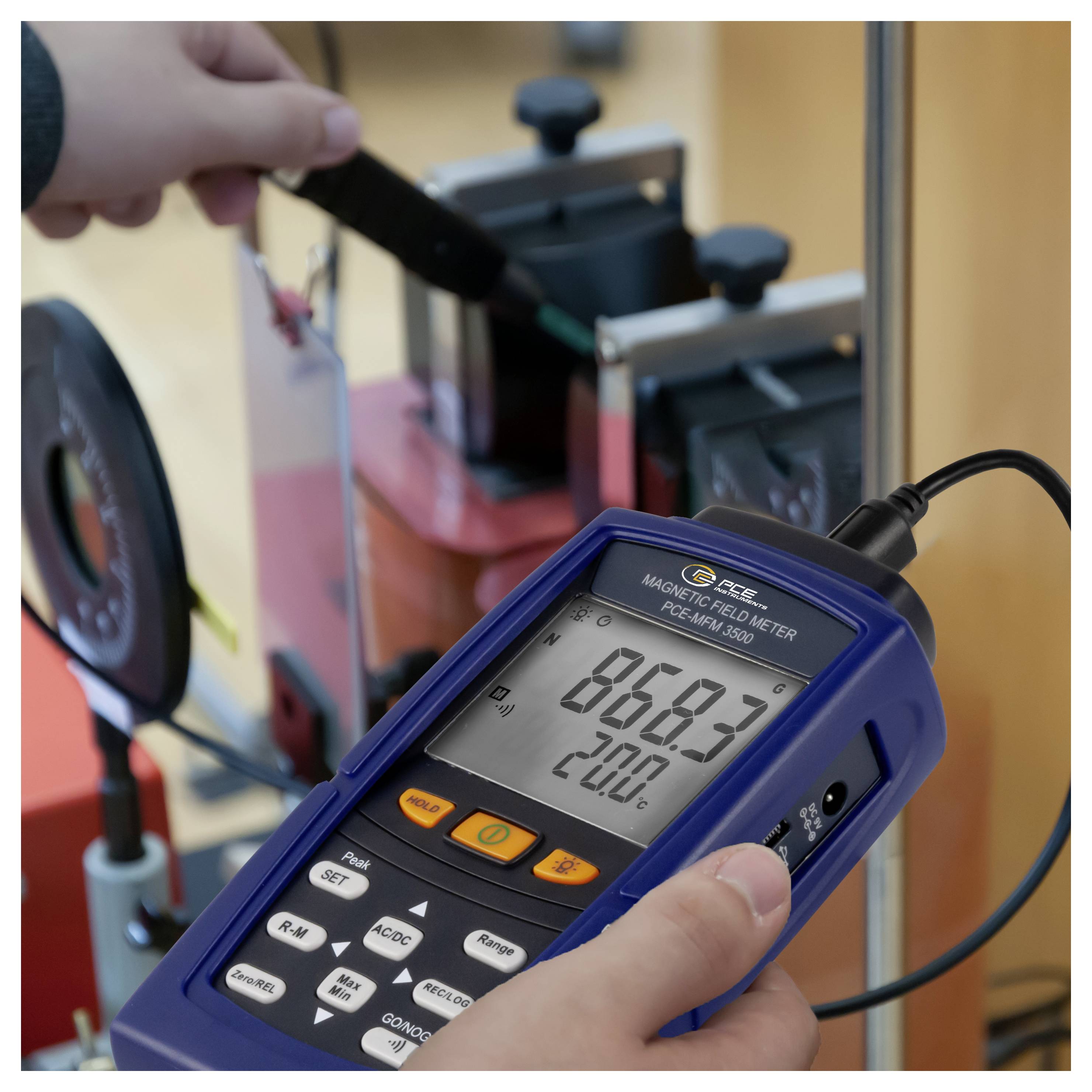 Person mit einem tragbaren Magnetfeldmessgerät in der Nähe von Laborausrüstung, die eine Anzeige von 868,3 und 20,0°C auf dem Bildschirm misst und anzeigt.