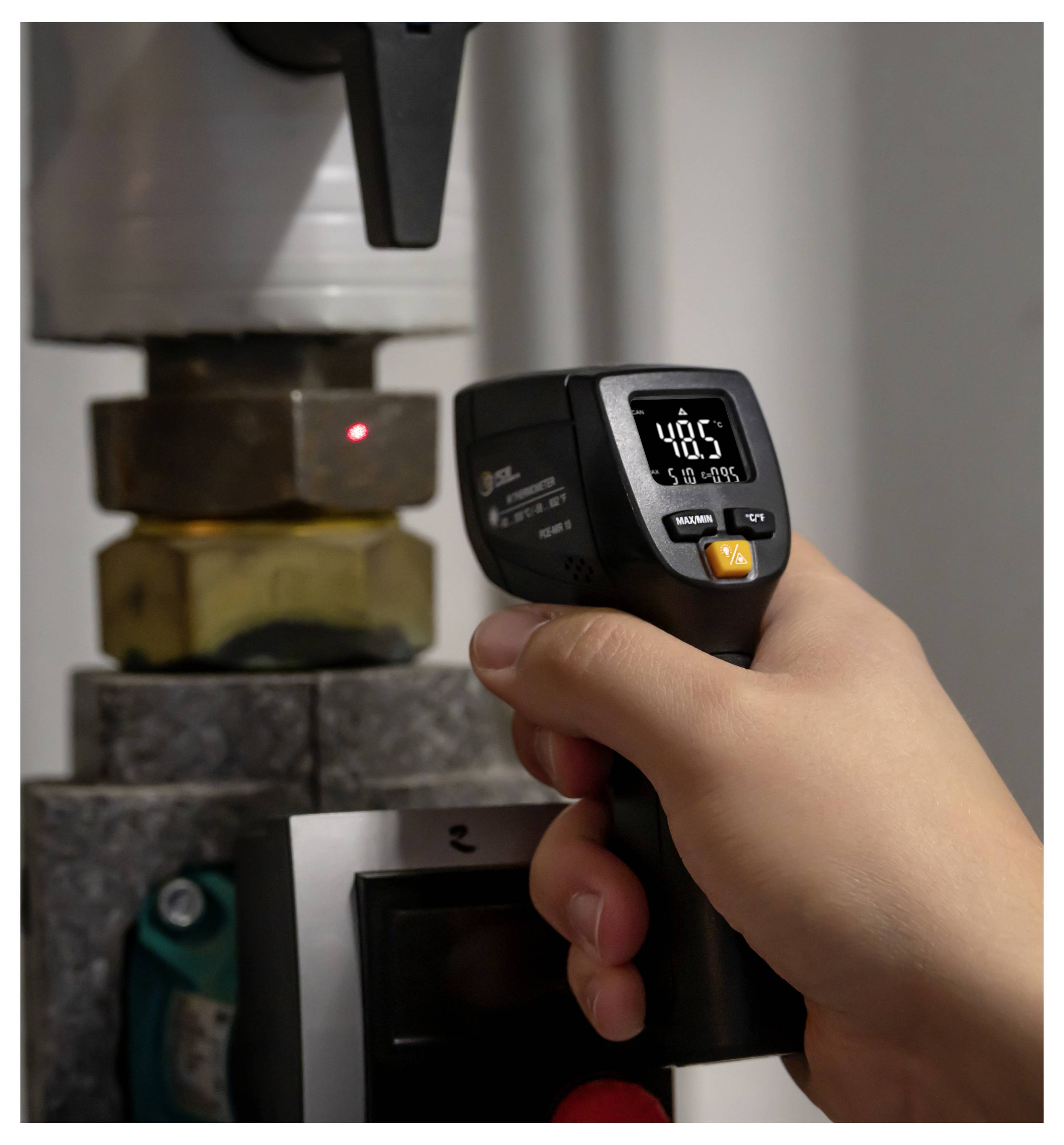 Eine Hand hält ein digitales Infrarot-Thermometer, das ein Metallteilmaschine misst und zeigt 48,5 auf dem Display an.