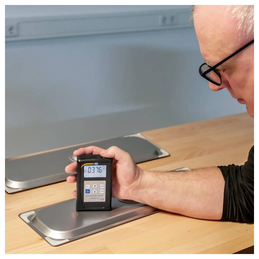 Ein Mann hält ein tragbares digitales Messinstrument über ein Metallpanel auf einem Holztisch und misst offenbar einen Oberflächenwert.